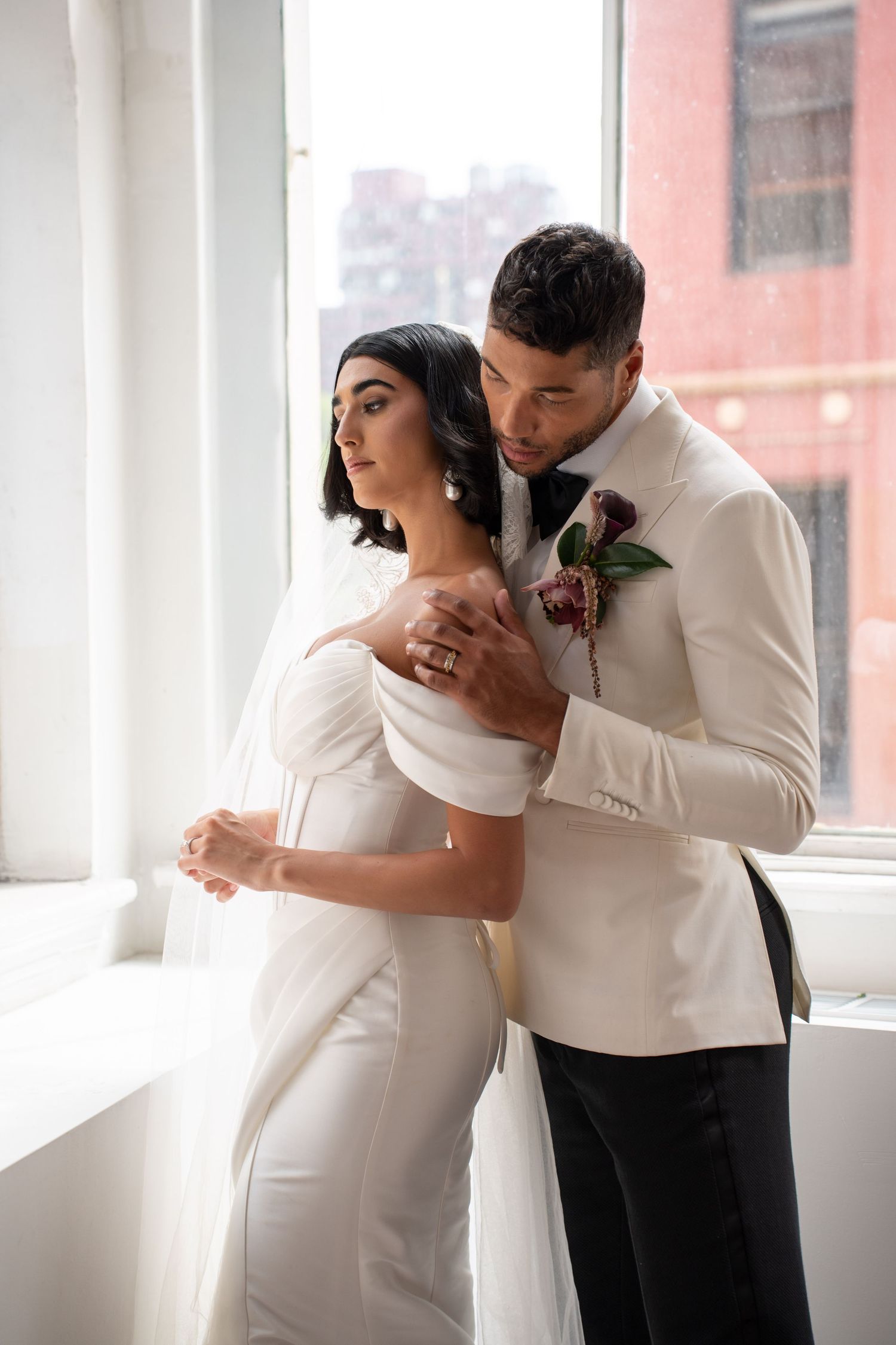 Groom in white tuxedo tenderly embracing bride in off-shoulder white gown by bright window.