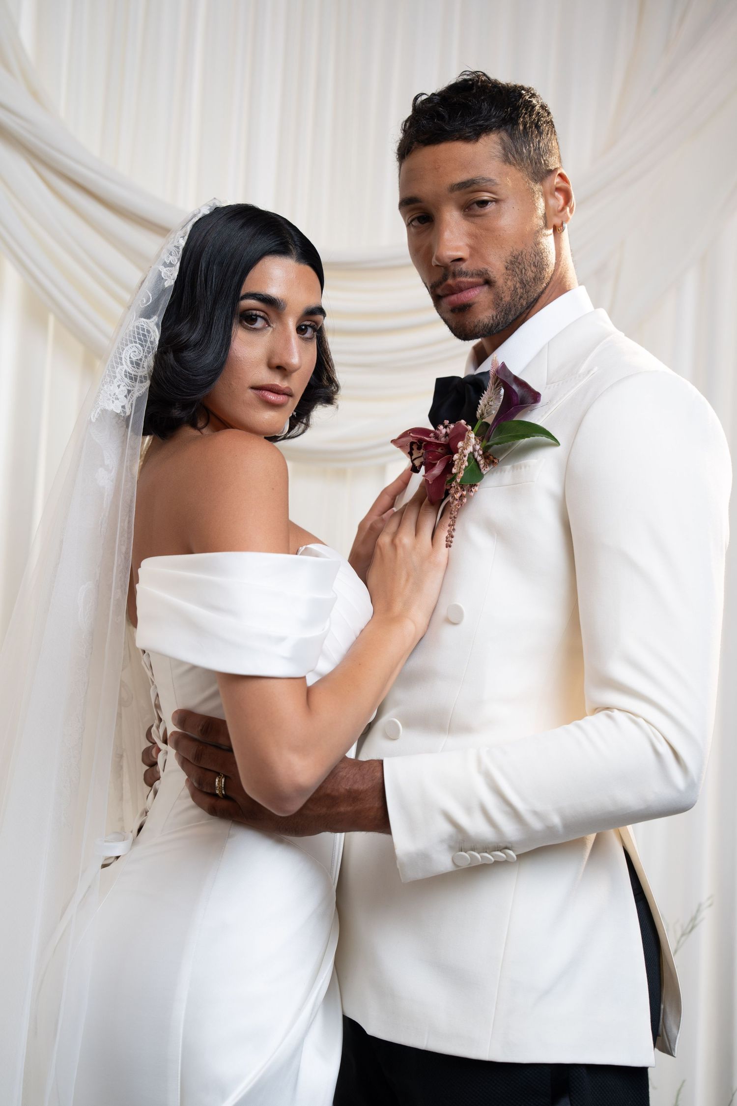 Bride in white off-shoulder gown adjusts grooms boutonniere while posing against draped white backdrop.