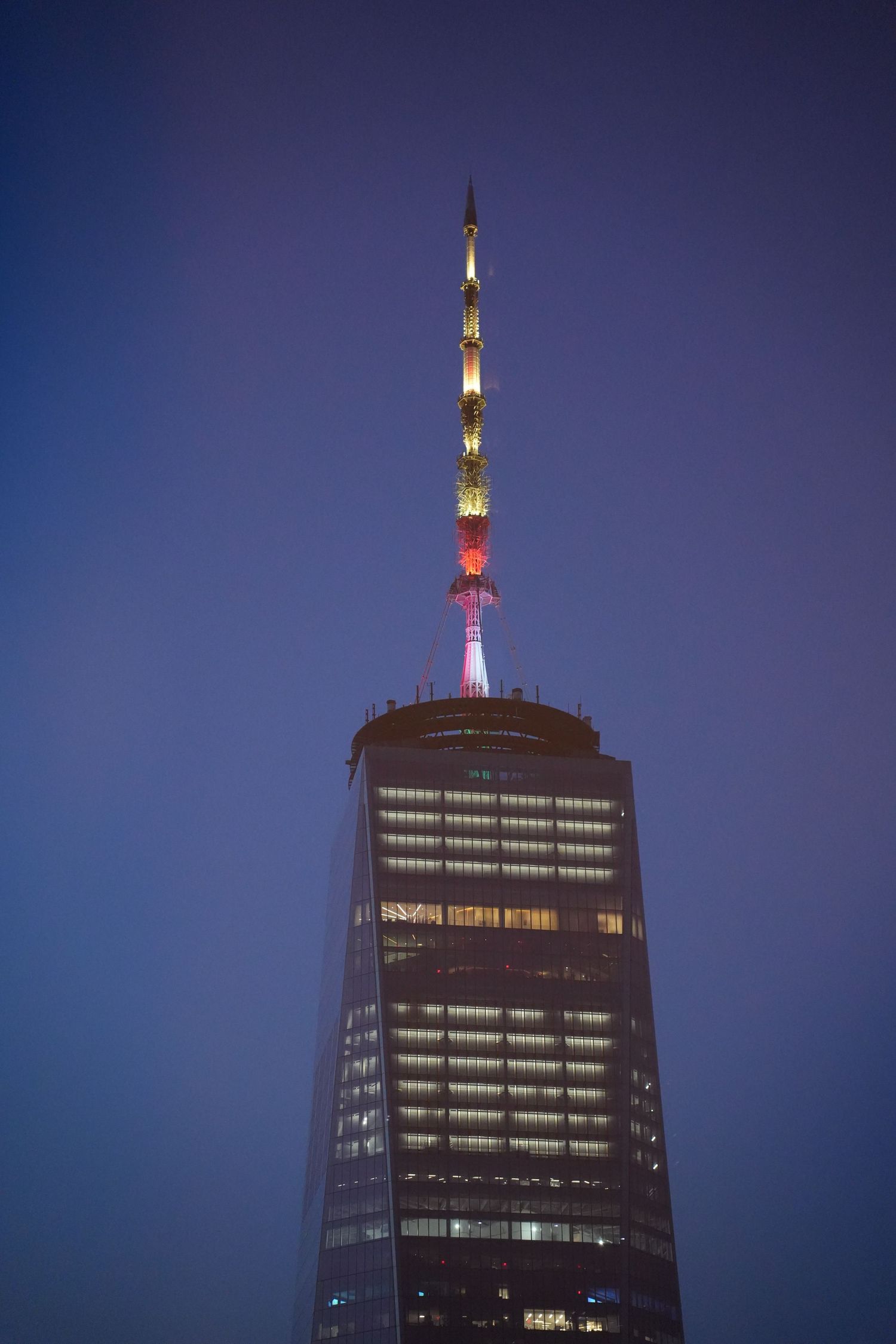 One World Trade Center illuminated at dusk with colorful antenna lights against a blue sky.