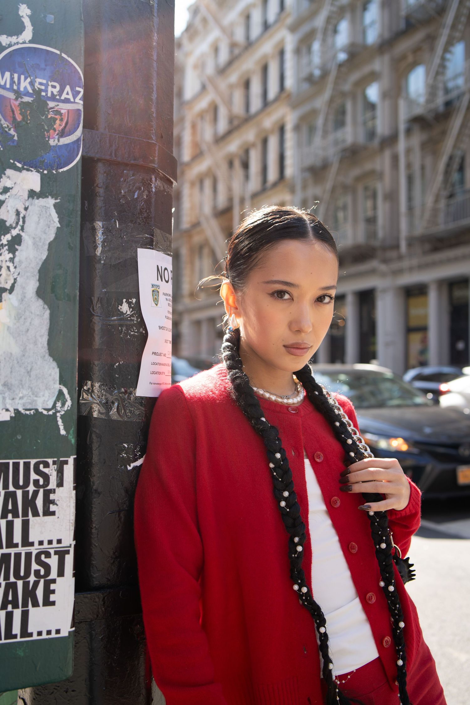 Stylish Asian woman in red cardigan with braided hair leans against graffiti pole on sunny NYC street.