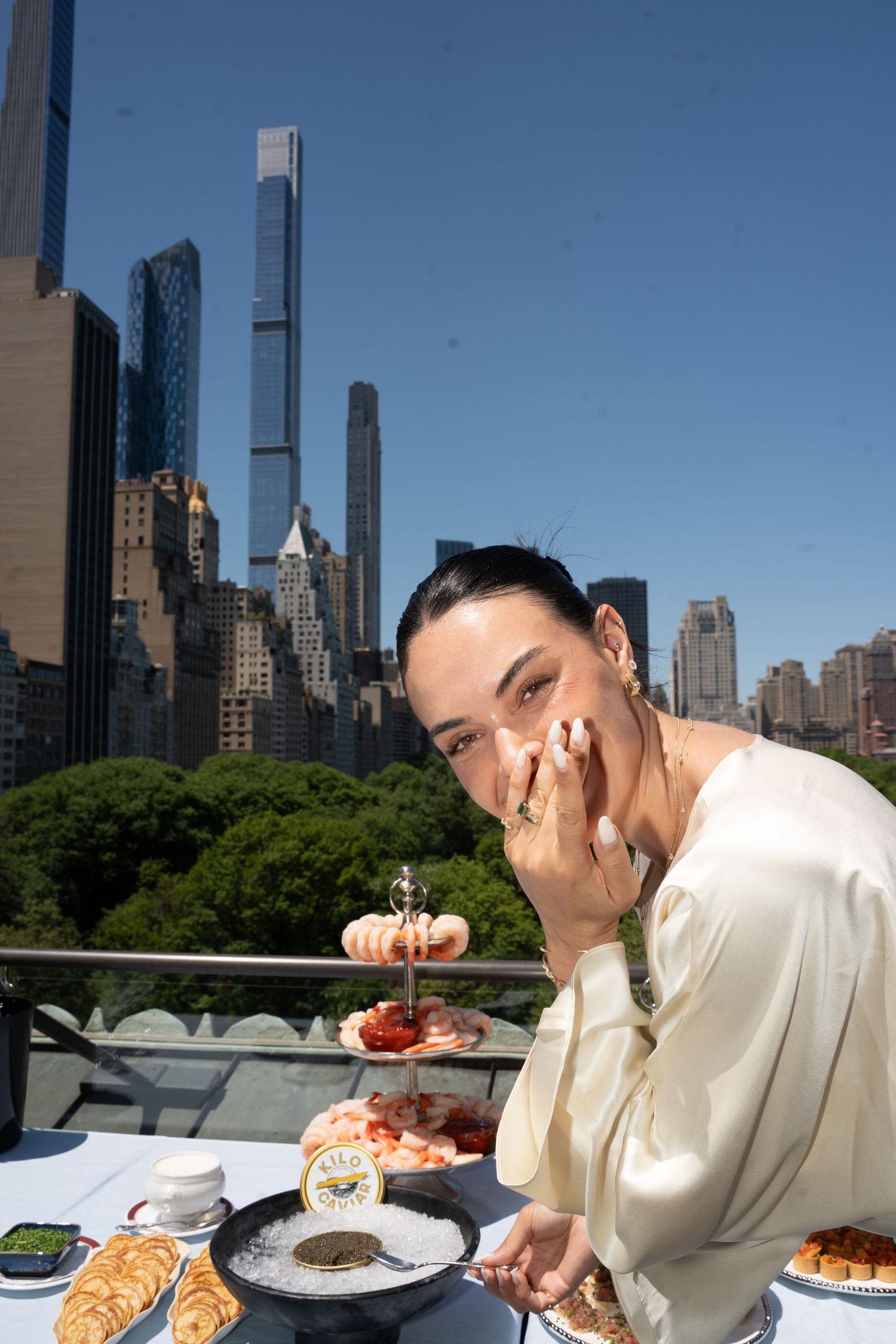 Woman smiling while enjoying seafood tower with caviar on NYC rooftop with Manhattan skyline view.