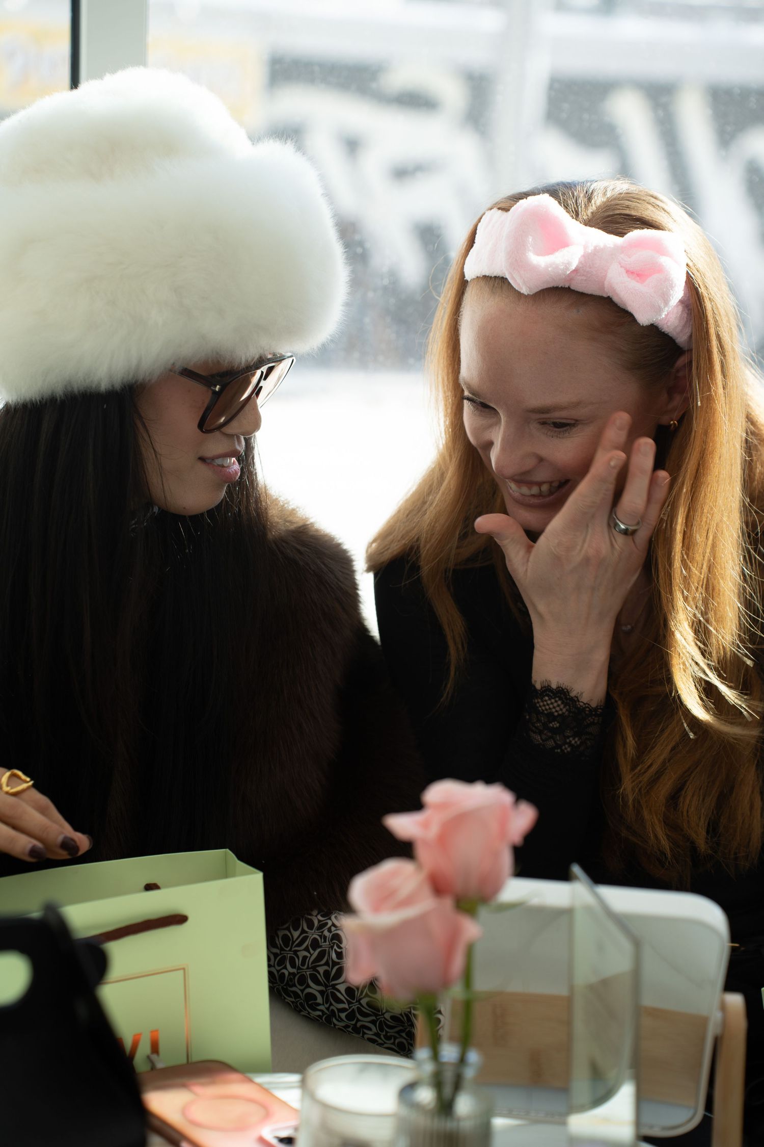 Two women smile together, one in a white fur hat and one with a pink bow headband.