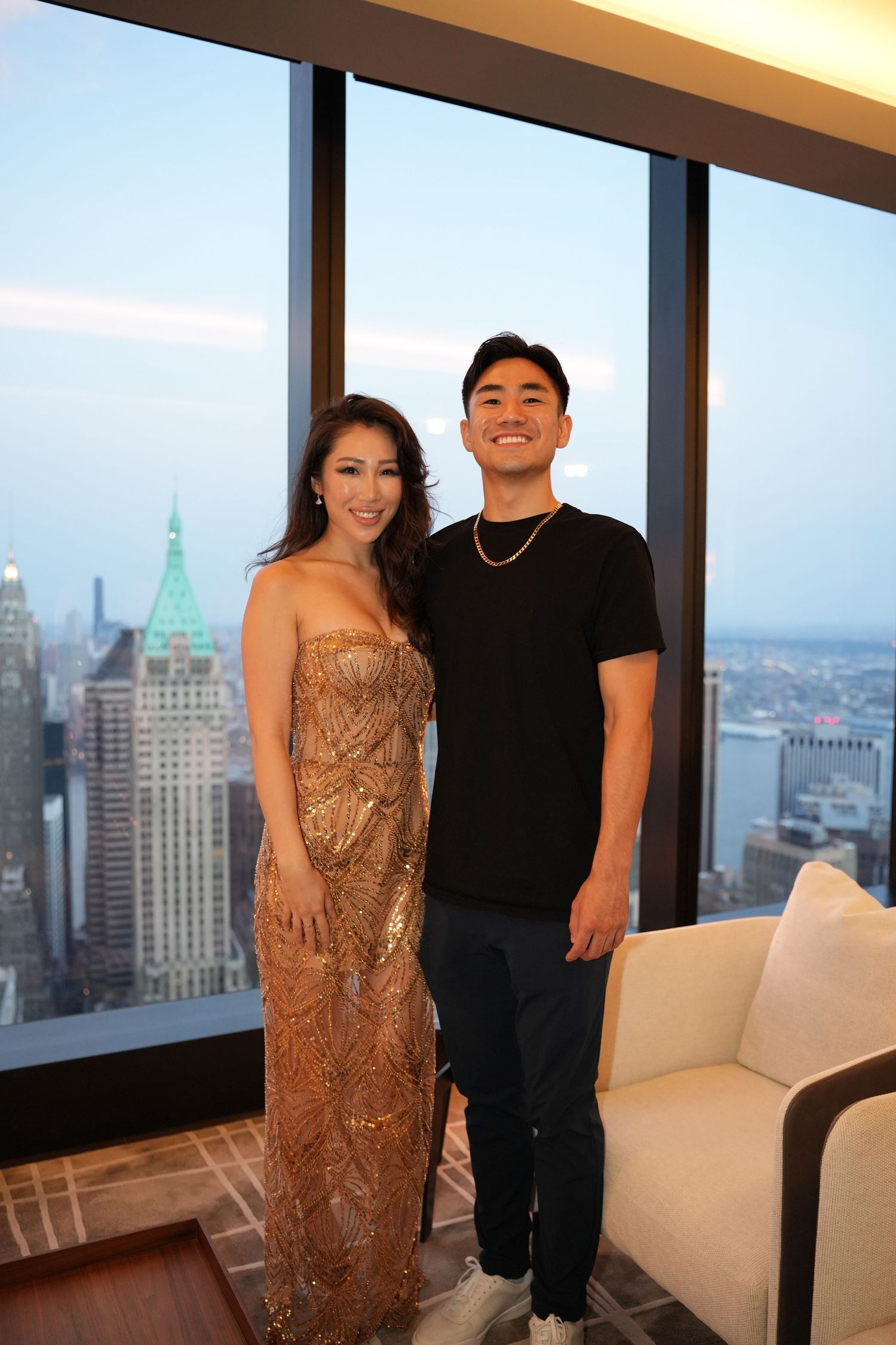 Couple posing in elegant attire with New York City skyline visible through floor-to-ceiling windows.