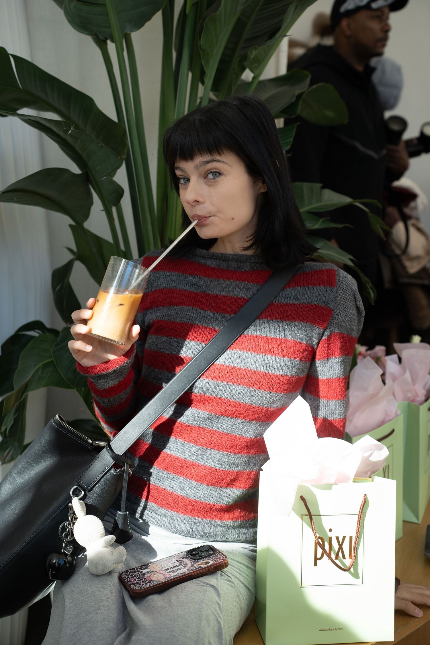 Woman in striped sweater drinking orange juice beside a green Pixi beauty shopping bag.