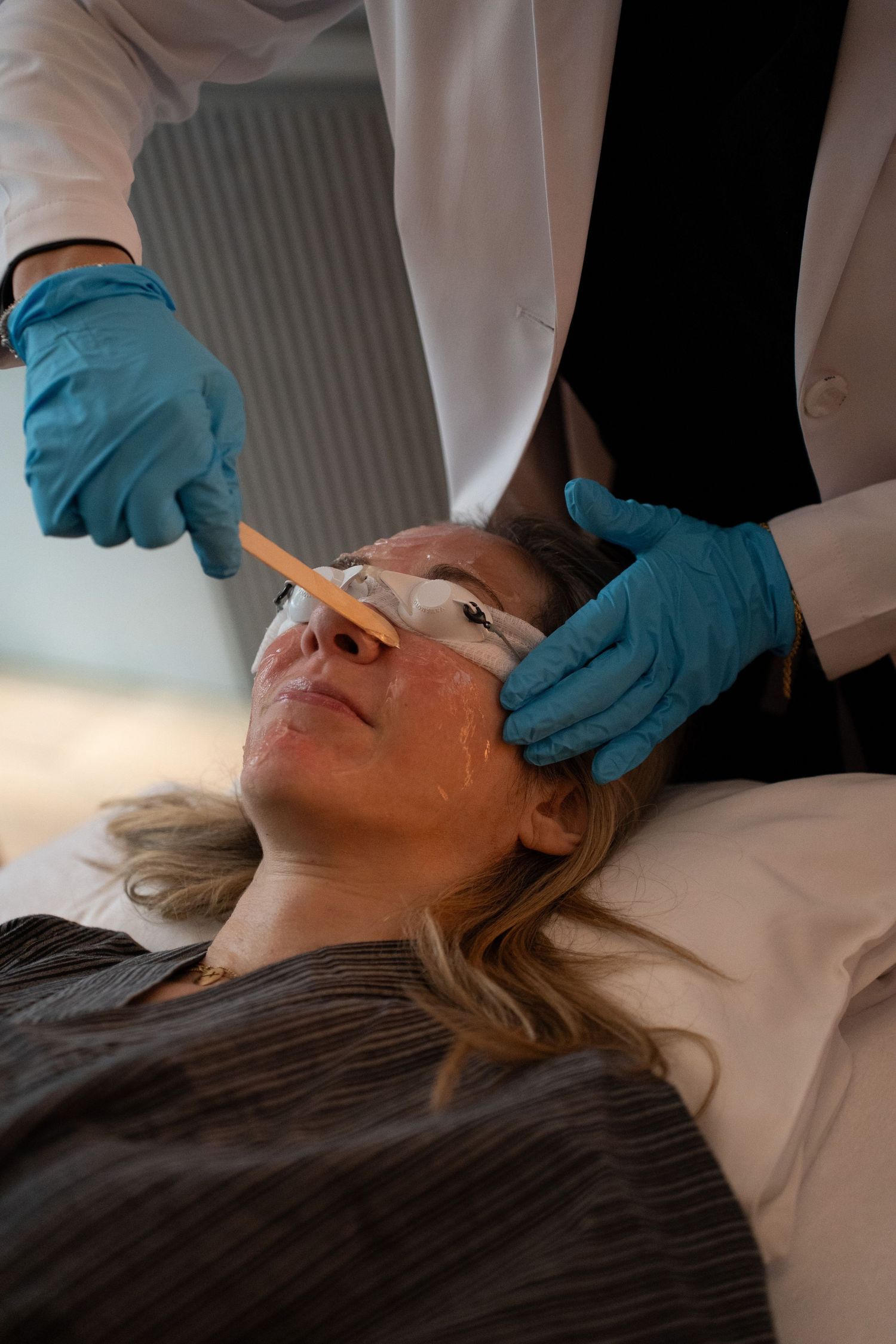 Medical professional in blue gloves applying treatment serum to female patient's face with a spatula.