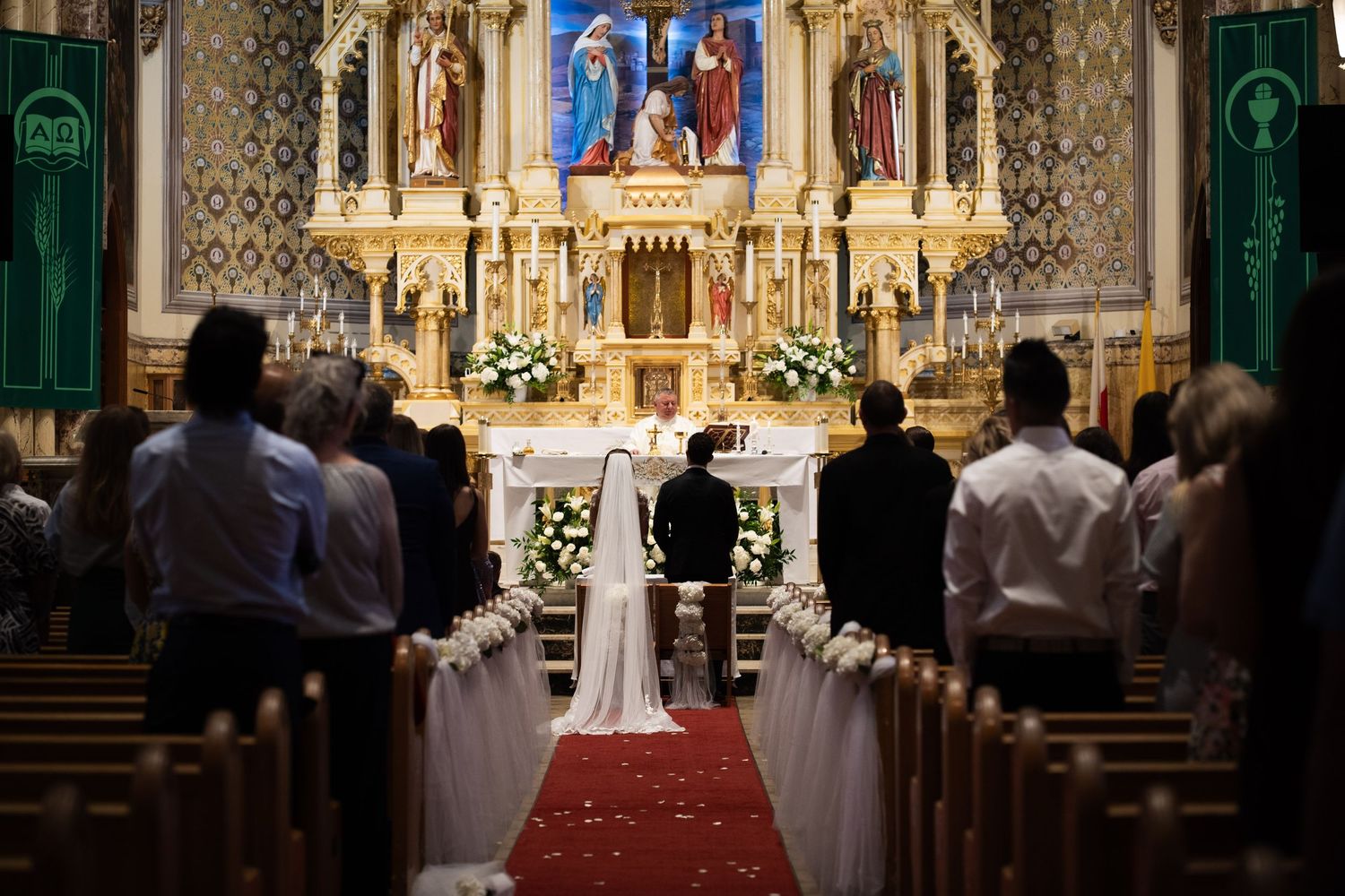 Wedding couple stands at altar of grand Catholic church with guests seated in pews.