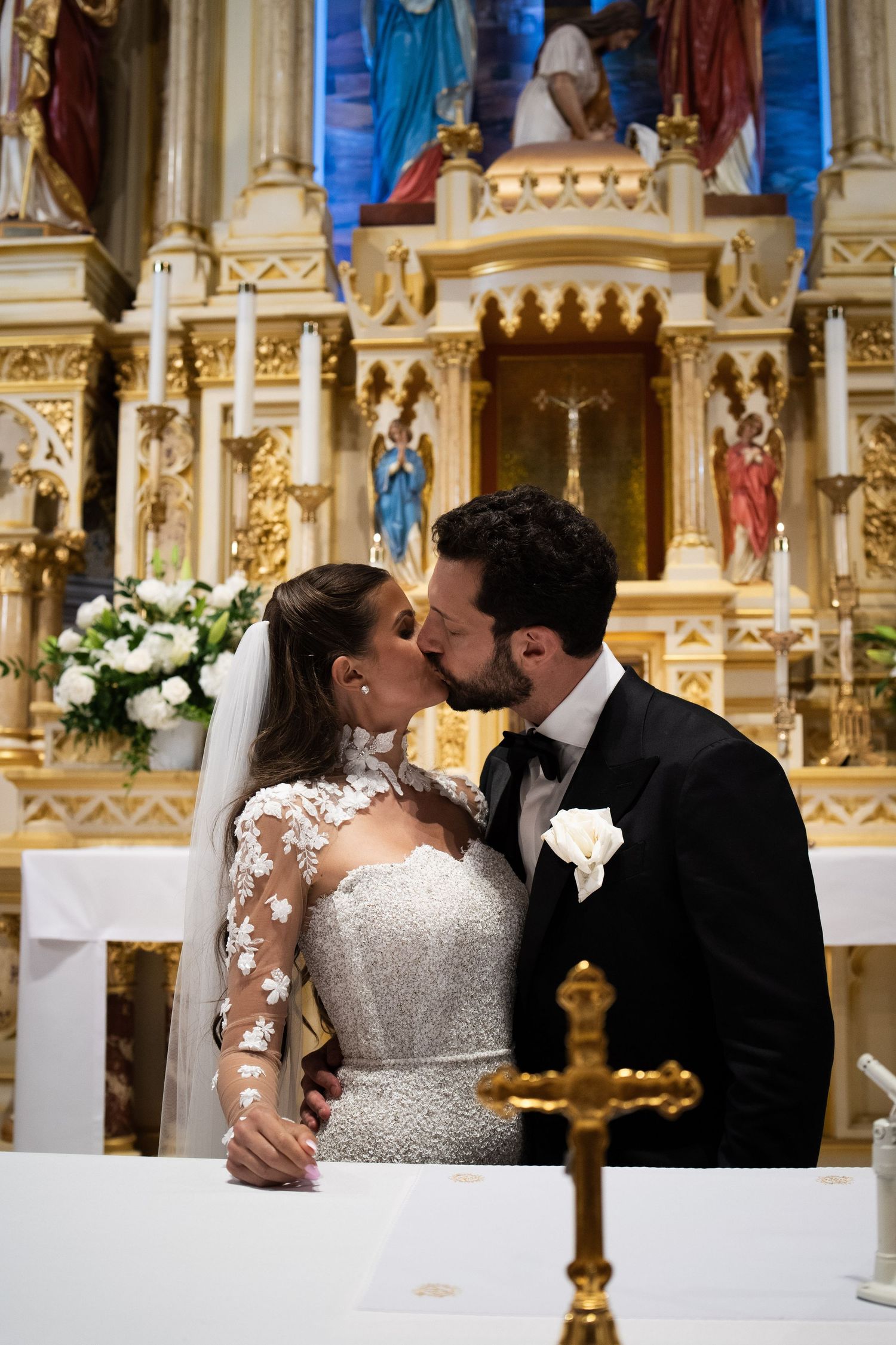 Newlyweds share first kiss at gilded church altar, bride in beaded lace gown with veil.