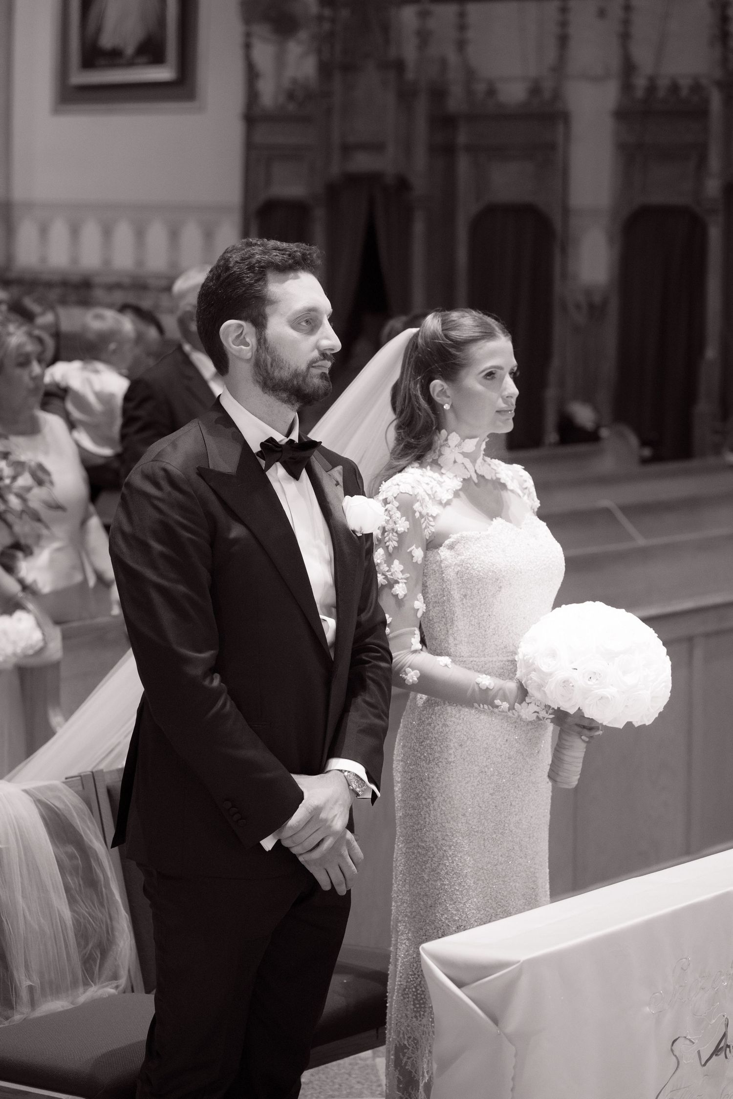 Bride in lace gown holding white bouquet and groom in tuxedo stand together during their church wedding ceremony.