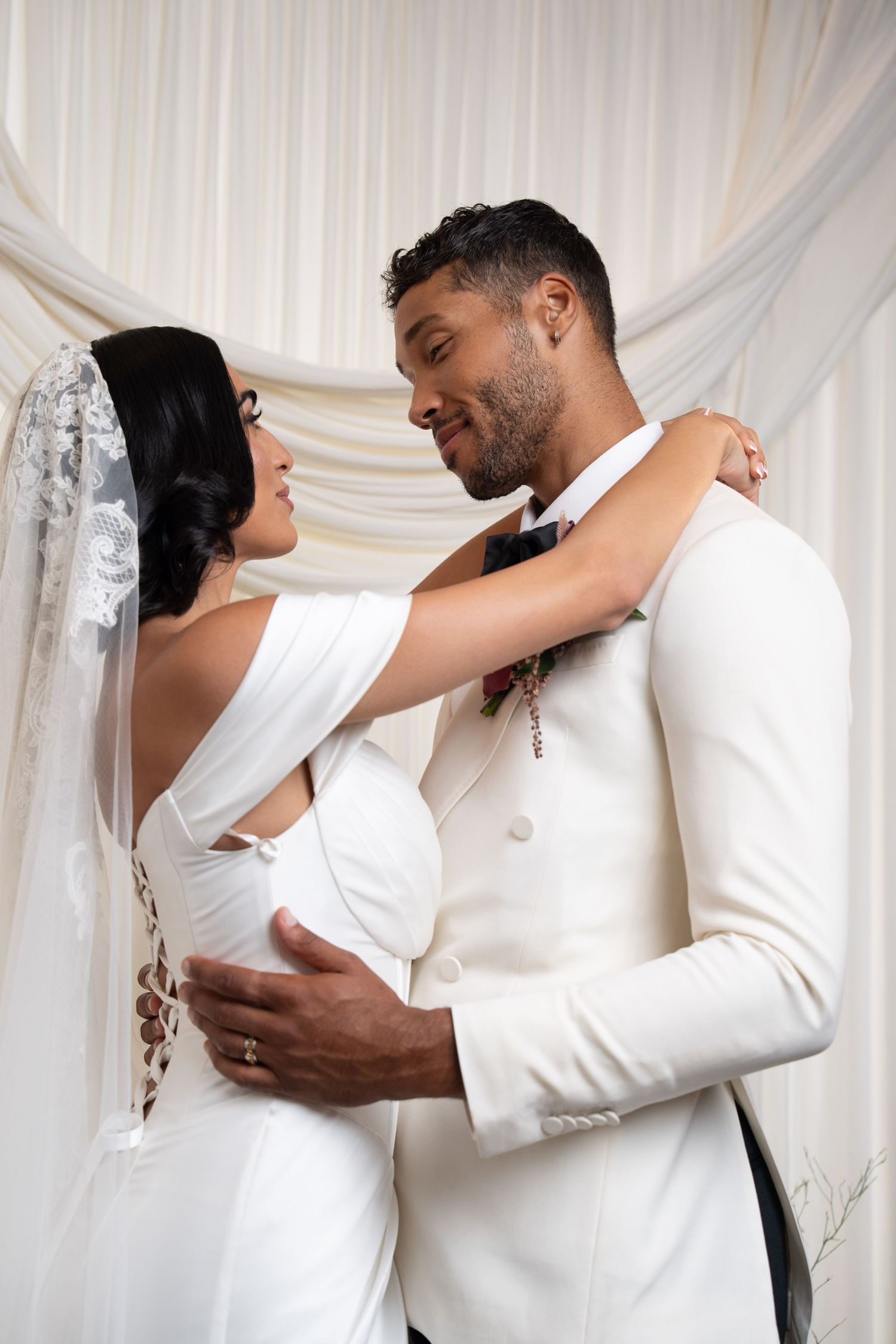 Bride and groom in white wedding attire embrace and smile at each other before draped backdrop.