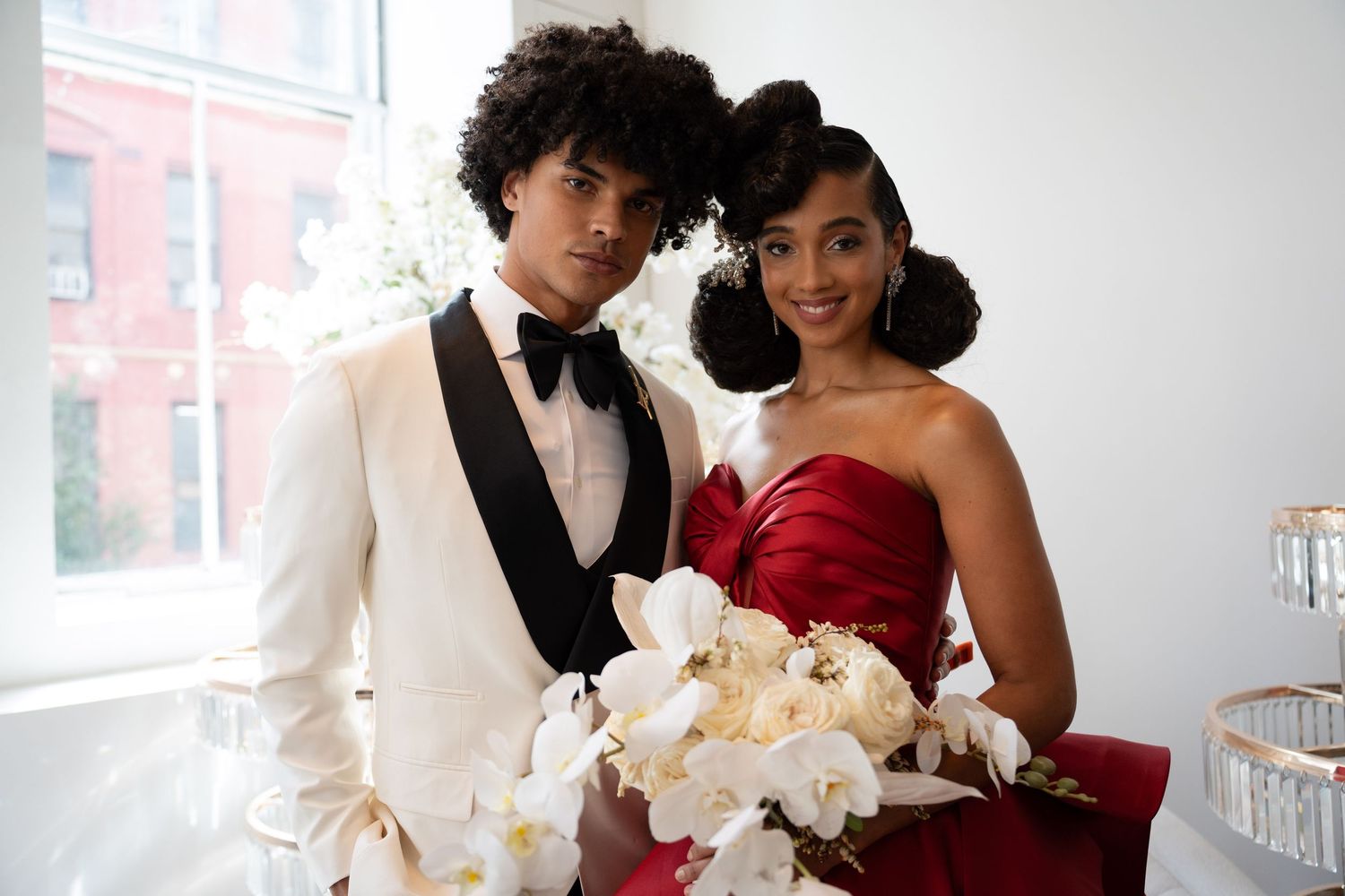 Young Black wedding couple smiling, groom in white tuxedo with bow tie, bride in red gown with floral bouquet.