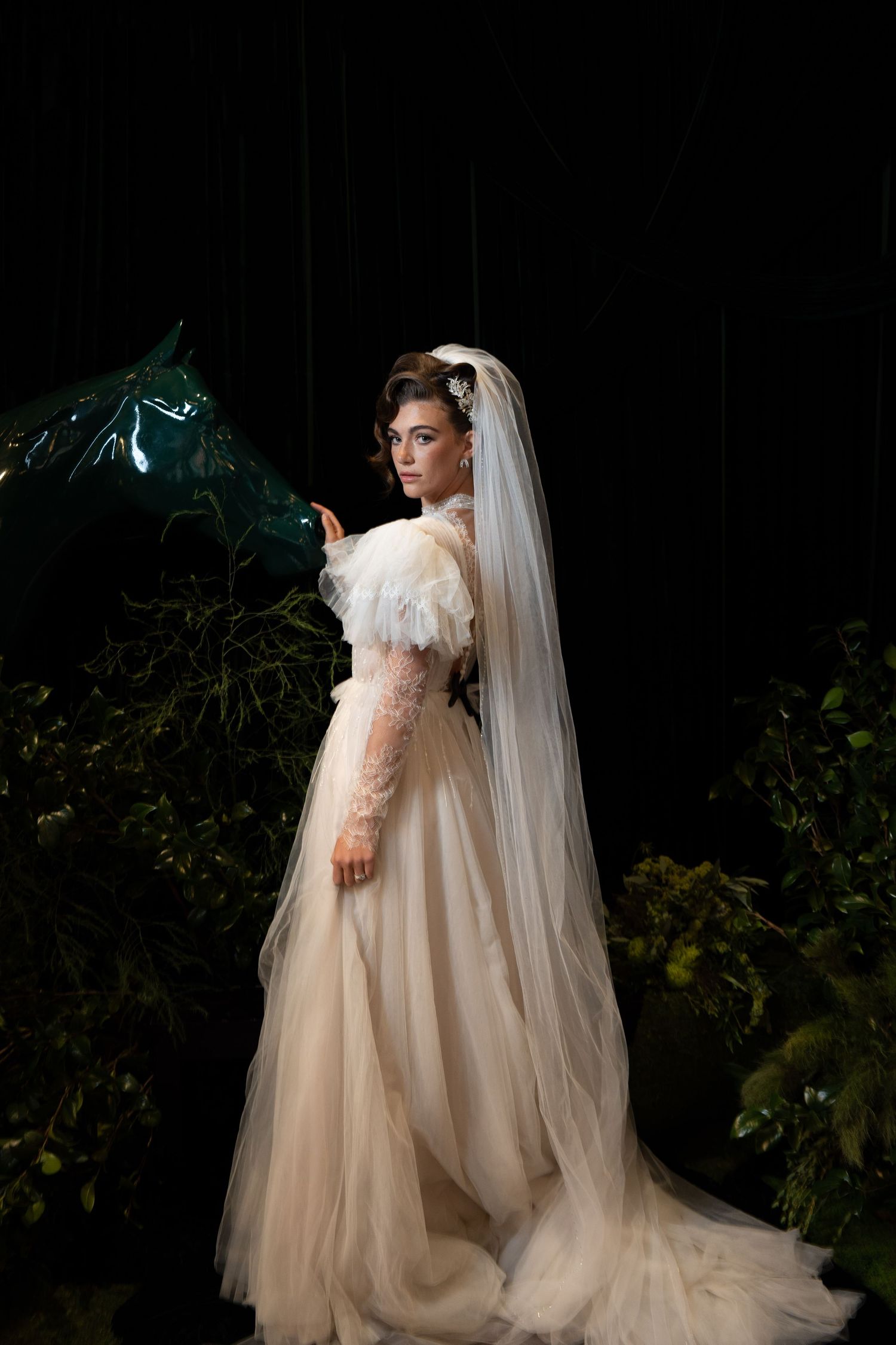 Bride in champagne tulle ball gown with cathedral veil stands gracefully among dark green plants.