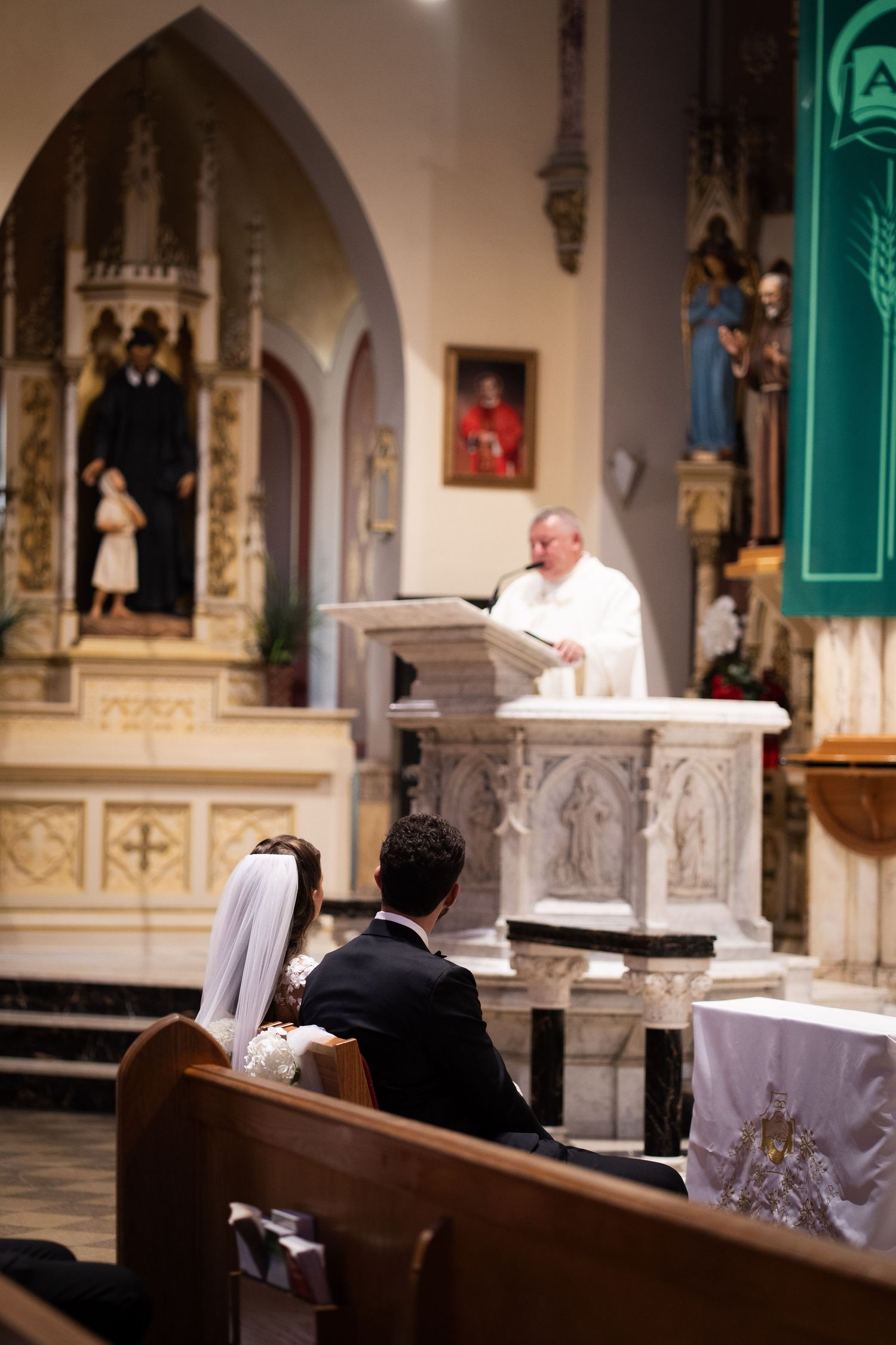 Bride and groom seated in church pew during Catholic wedding ceremony with priest reading at pulpit.