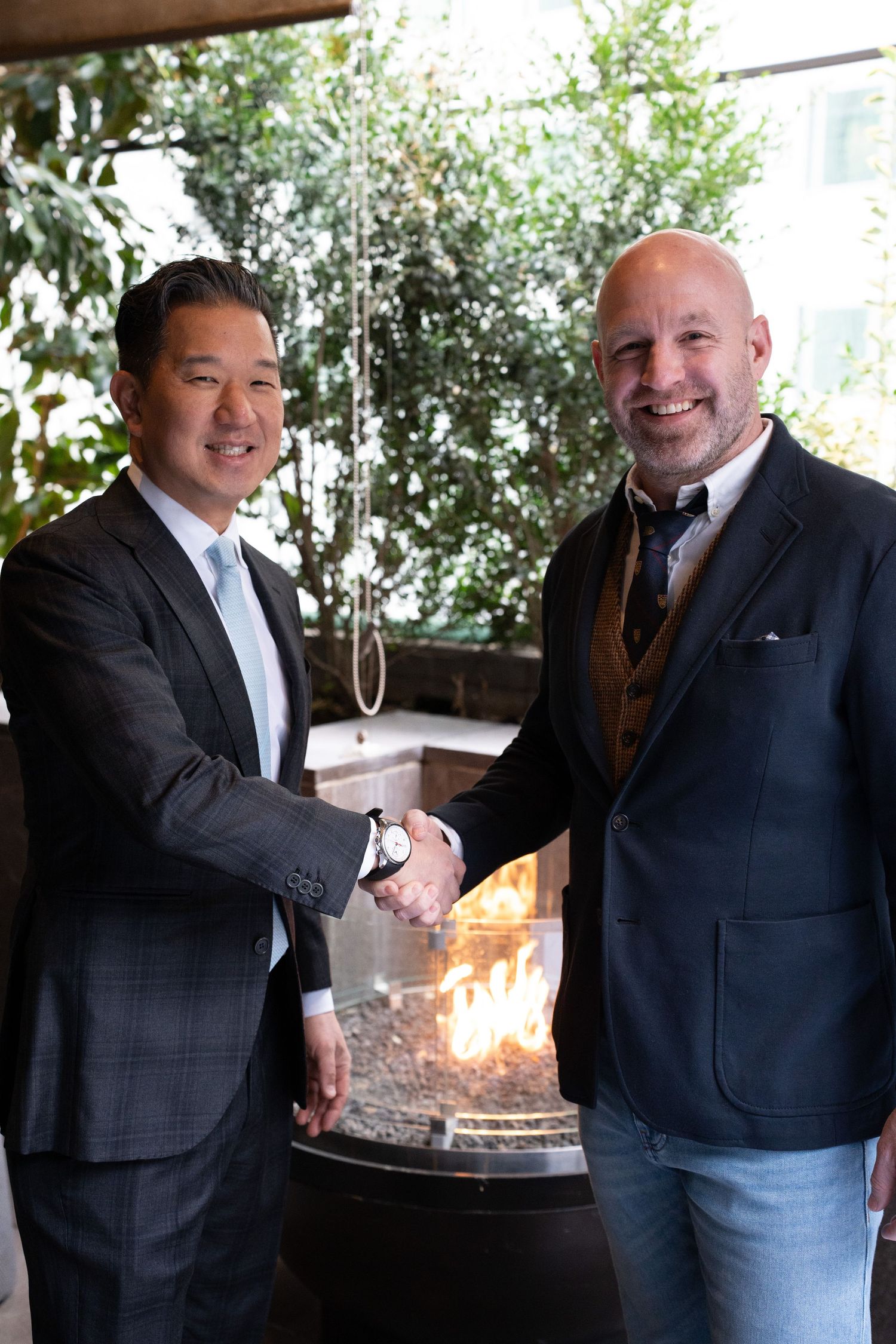 Two professional men shaking hands in front of an outdoor fire feature at a business event.