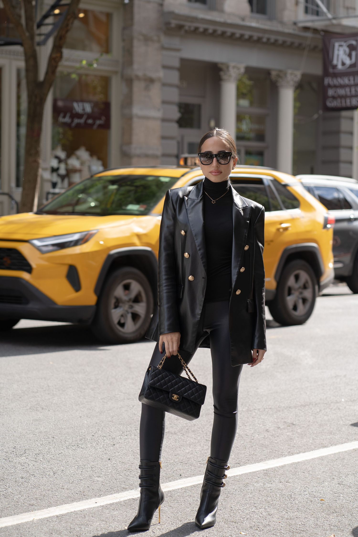 Woman in all-black leather blazer, leggings, and heeled boots holding Chanel bag with yellow taxi in background.