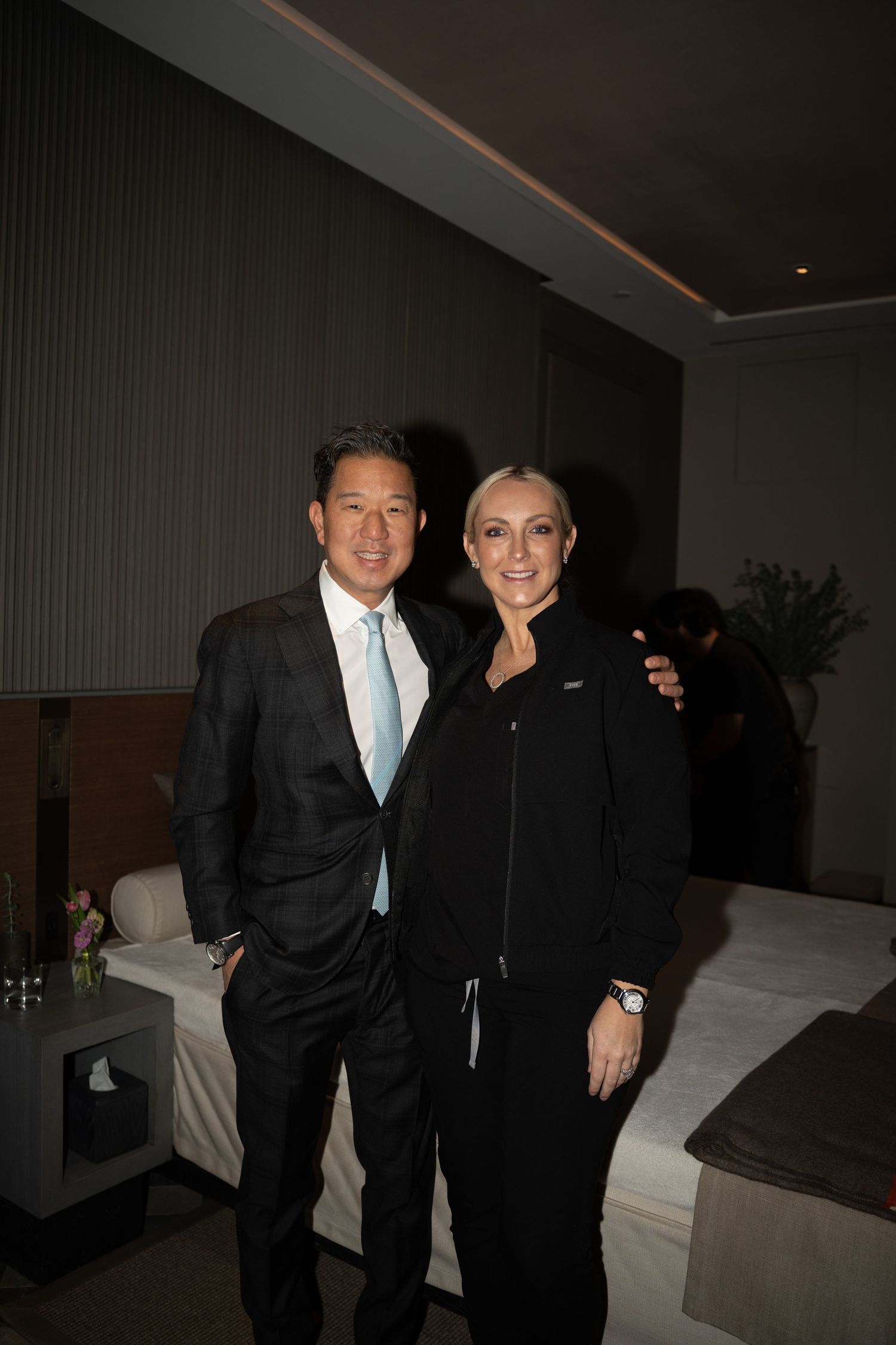 Asian man in dark suit and blonde woman in black uniform posing together at a medical spa event.