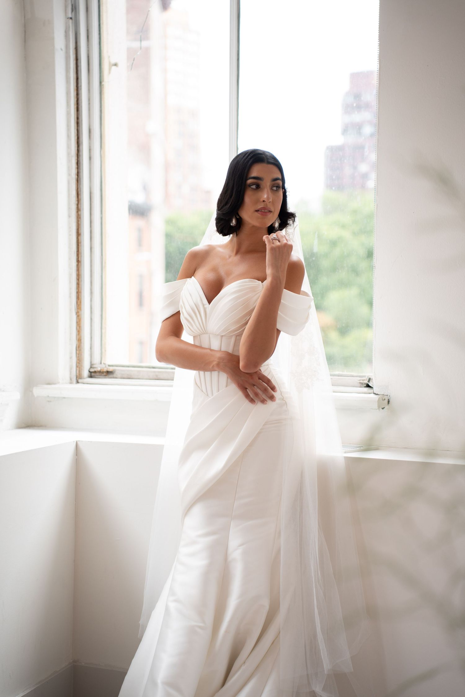 Elegant bride in off-shoulder white gown with draped veil standing by large bright window.