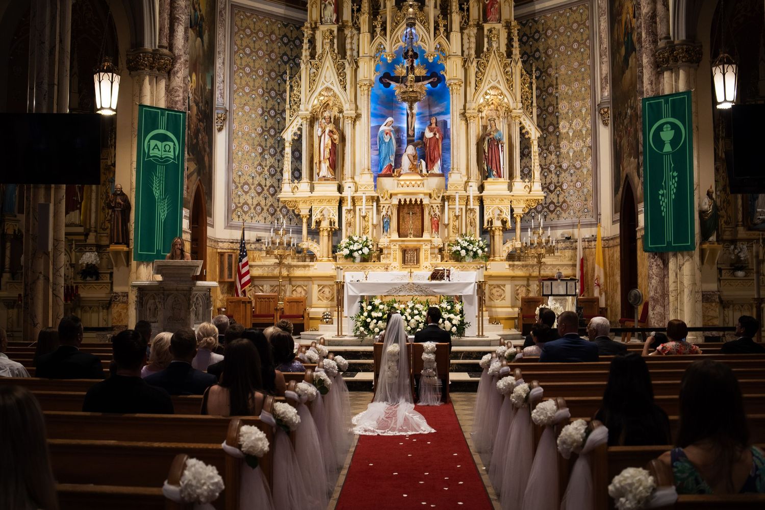 Wide view of ornate Catholic church wedding ceremony with couple at altar and guests in decorated pews.