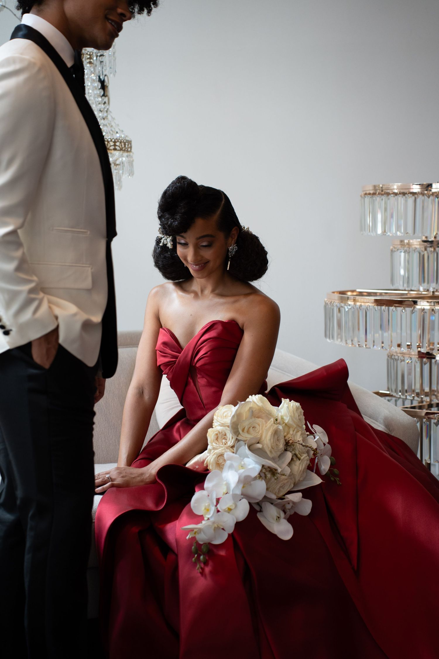 Elegant bride in red strapless gown admiring her white rose and orchid bouquet beside her groom at wedding.