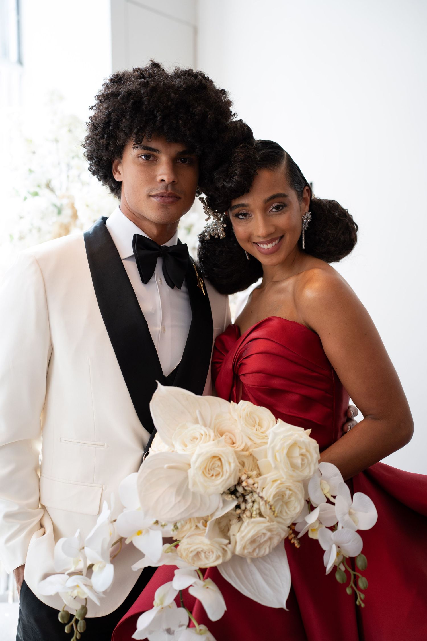 Elegant couple posing at wedding, man in white tuxedo with bow tie, woman in red strapless gown holding white floral bouquet.