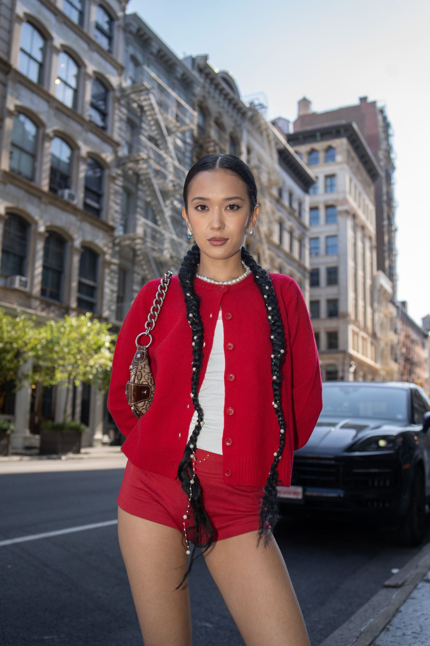 Young Asian woman in red cardigan outfit poses on NYC street with braided hair and designer handbag.