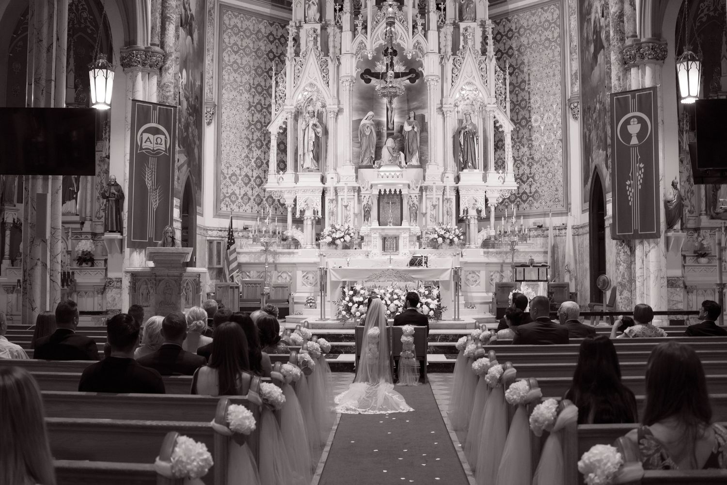 Wide-angle black and white view of a wedding ceremony in a grand Catholic church with ornate altar and pew decorations.