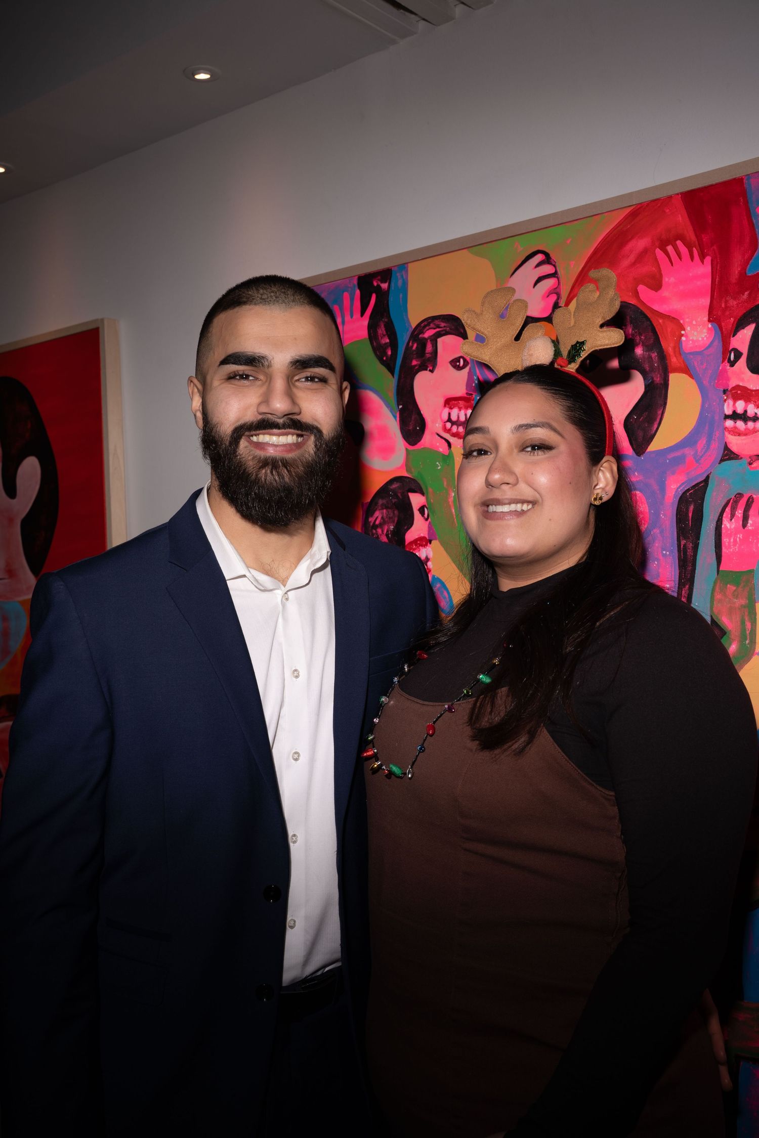 Man in blue blazer and woman in brown top smiling in front of vibrant colorful artwork at gallery.