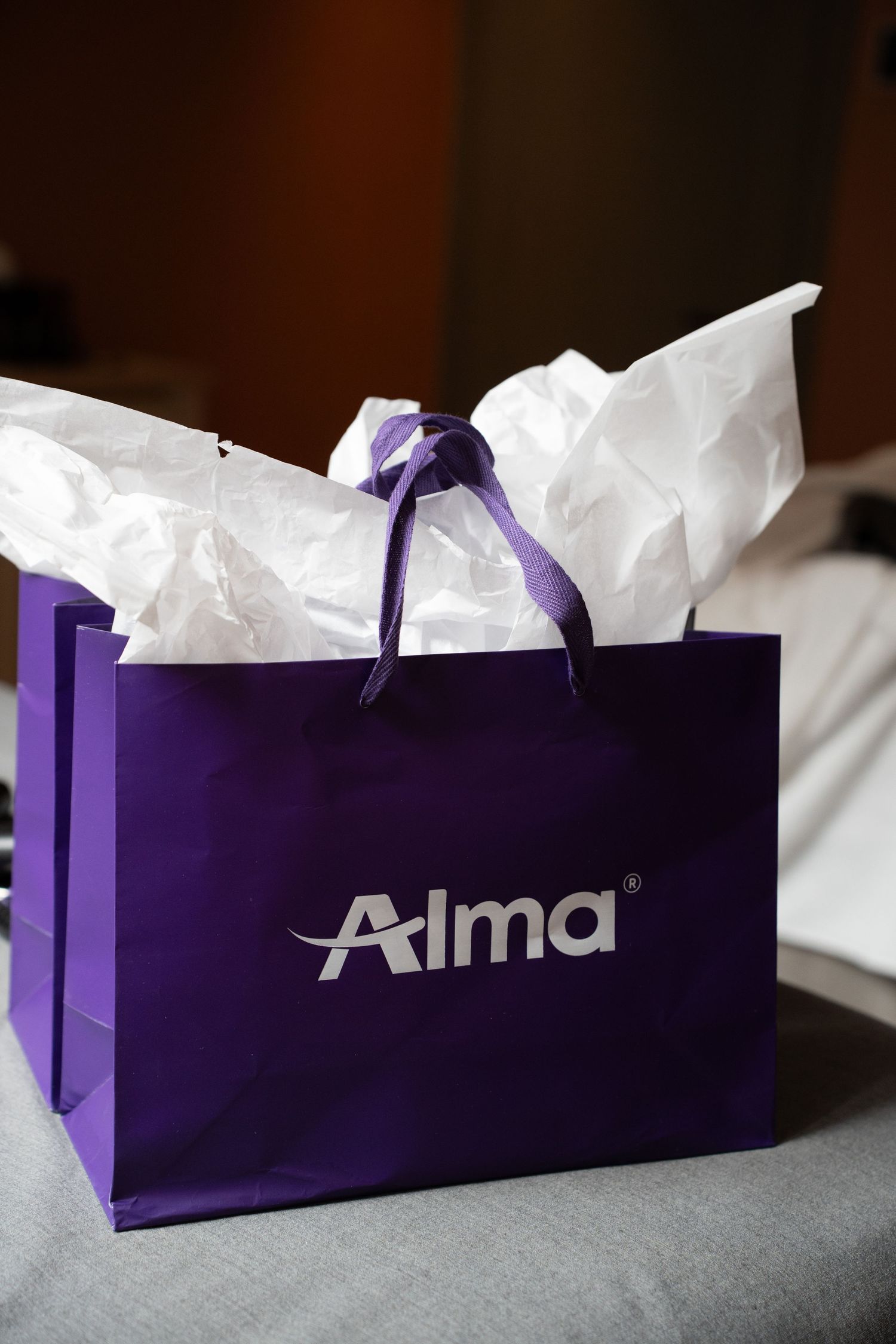 Purple Alma laser aesthetics branded gift bag with white tissue paper on a table.