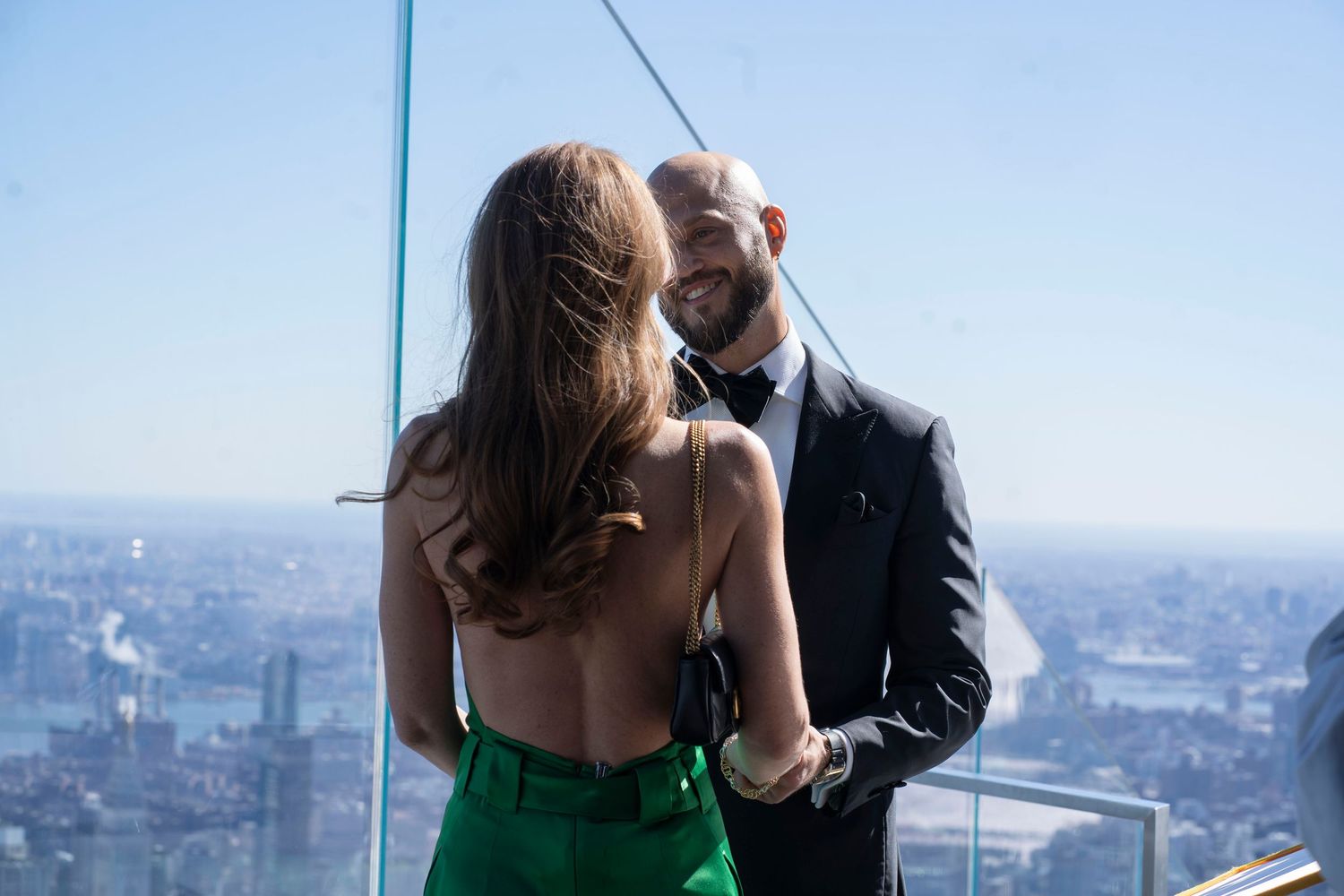 Couple embracing on rooftop with New York City skyline, woman in green dress, man in tuxedo.