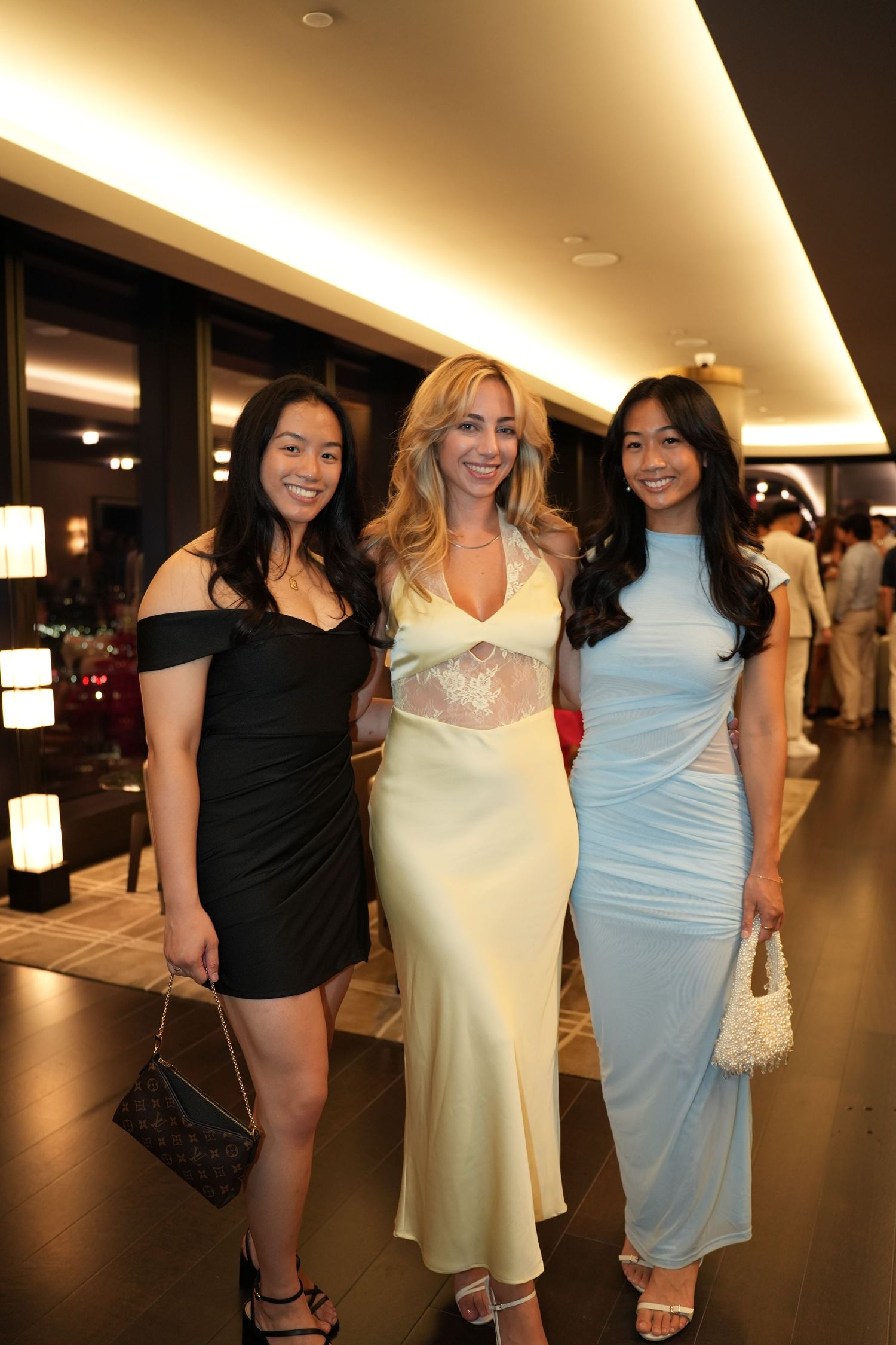 Three women in elegant dresses smiling together at an upscale evening social event.
