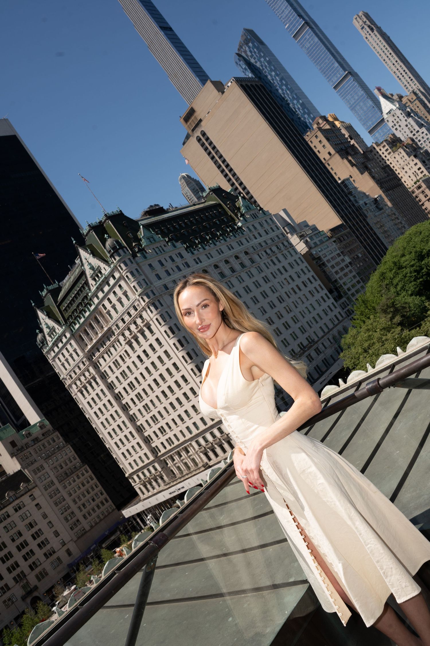 Blonde woman in white dress posing on rooftop terrace with NYC skyline and Plaza Hotel behind her.