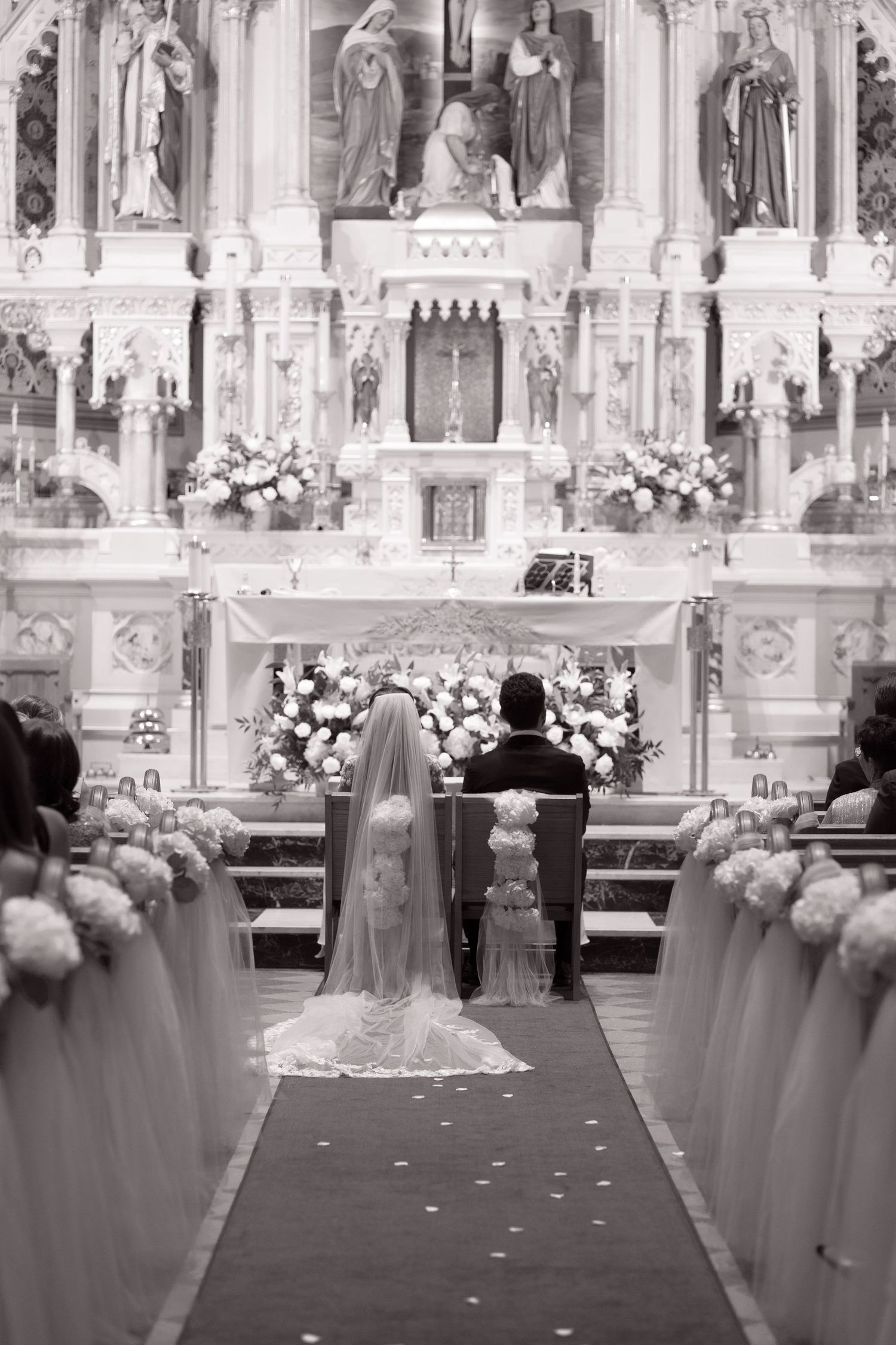 Bride in cathedral veil and groom sit before an ornate white altar decorated with large floral arrangements in a Catholic church.