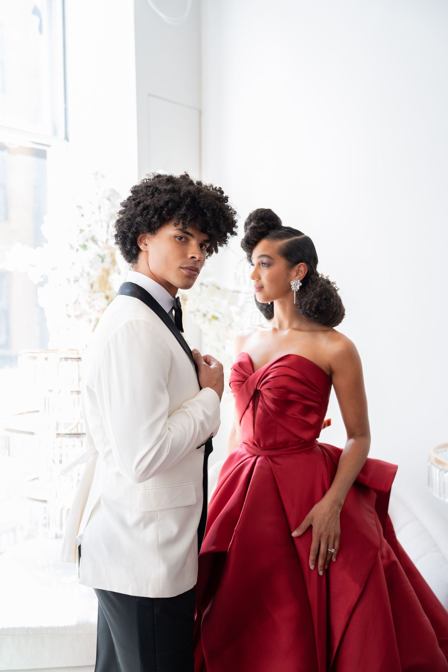 Elegant couple in white tuxedo and red strapless gown posing in bright wedding venue.