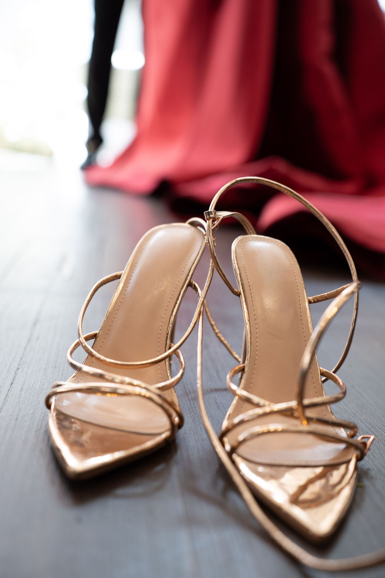 Gold strappy heeled sandals on dark floor with red gown and tuxedo visible in soft focus background.