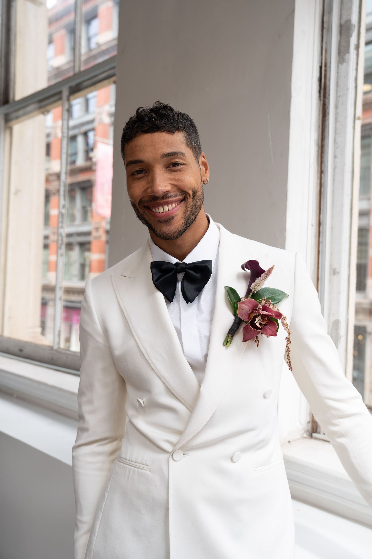 Smiling groom in white double-breasted tuxedo with black bow tie and burgundy floral boutonniere by window.