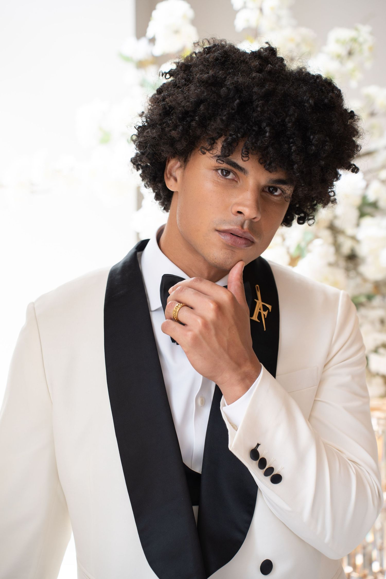 Elegant groom in white tuxedo jacket with black bow tie and gold brooch posing thoughtfully at wedding.