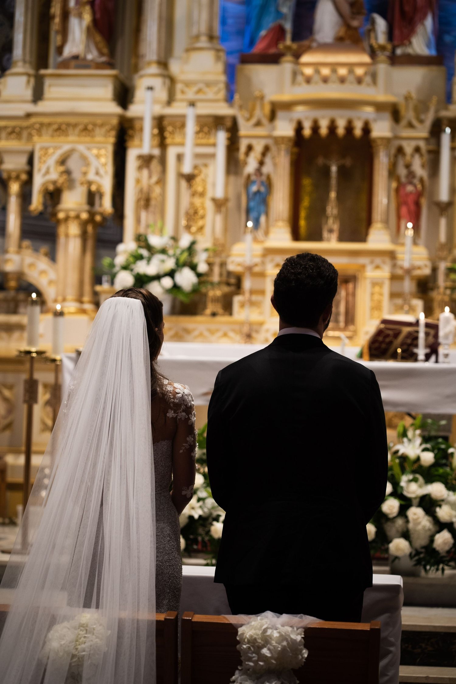 Bride in lace gown and groom in tuxedo kneel at church altar during Catholic wedding.