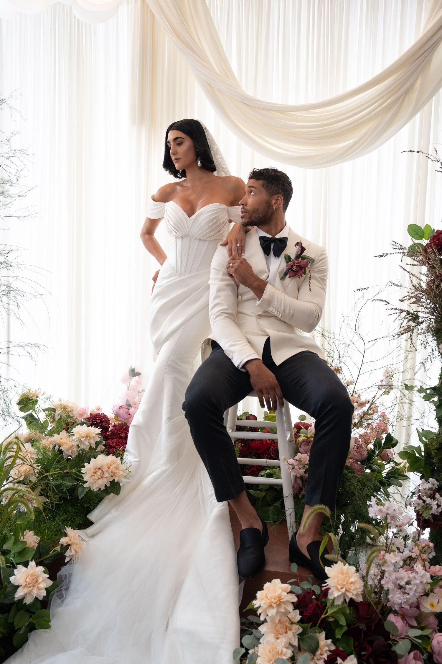 Bride in off-shoulder mermaid gown stands above seated groom in white tuxedo amid lush floral arrangements.