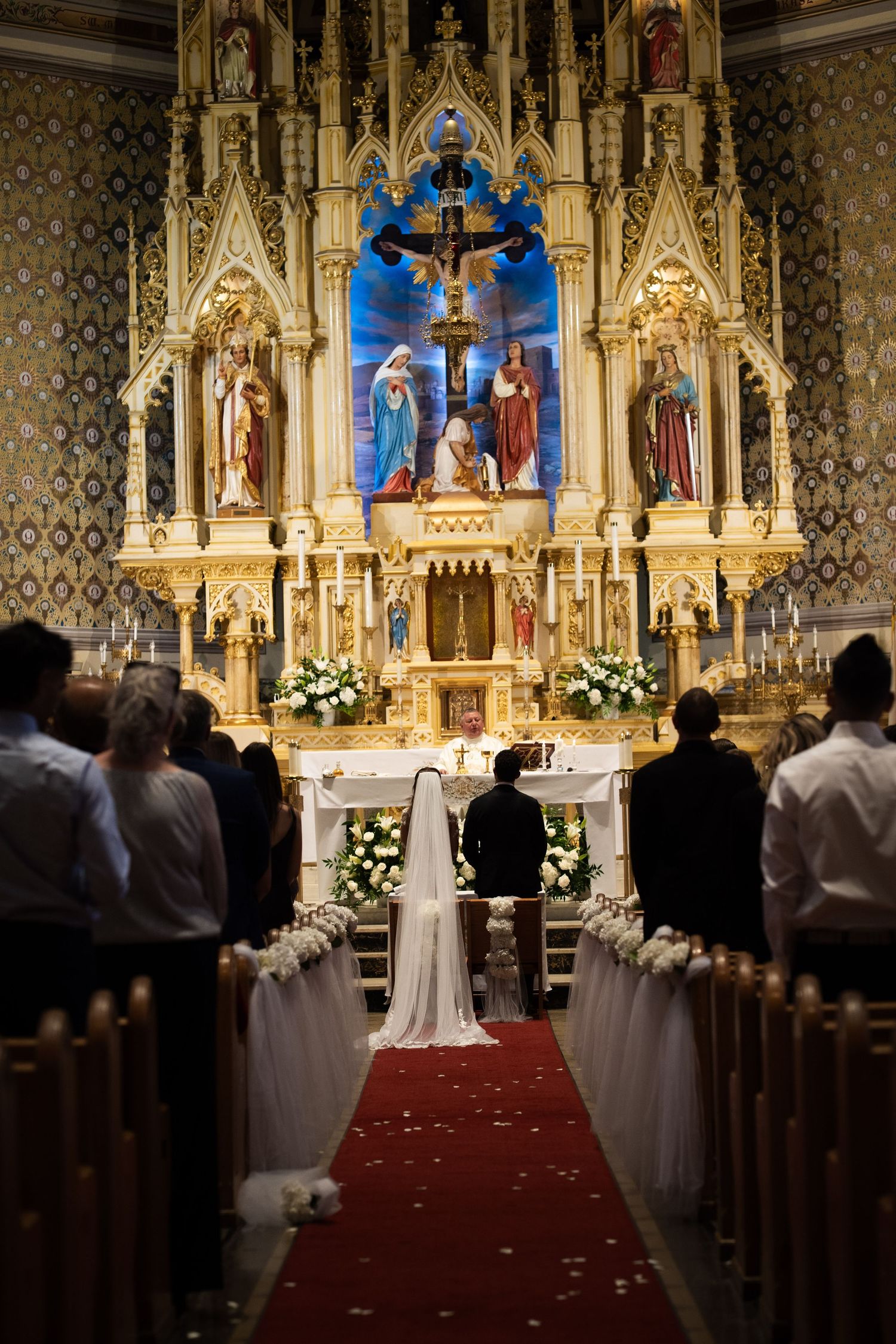 Bride and groom at ornate church altar with golden reredos and rose petal lined aisle.