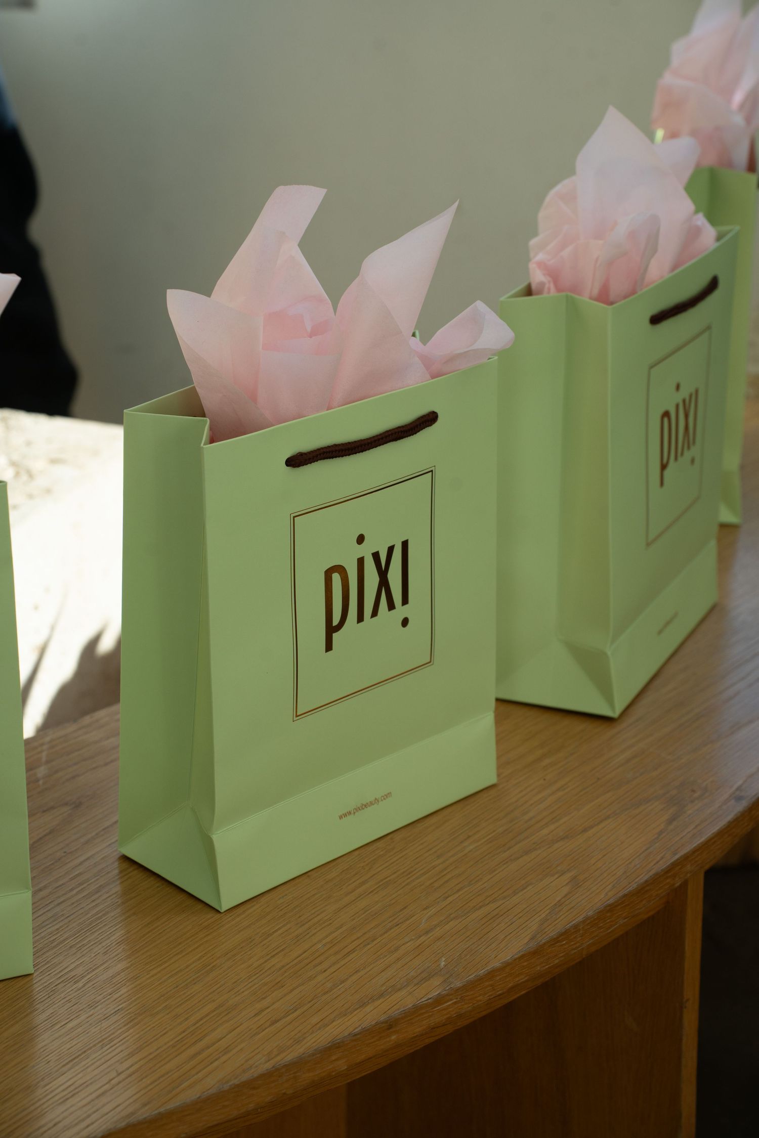 Two mint green Pixi beauty gift bags with pink tissue paper displayed on a wooden table.
