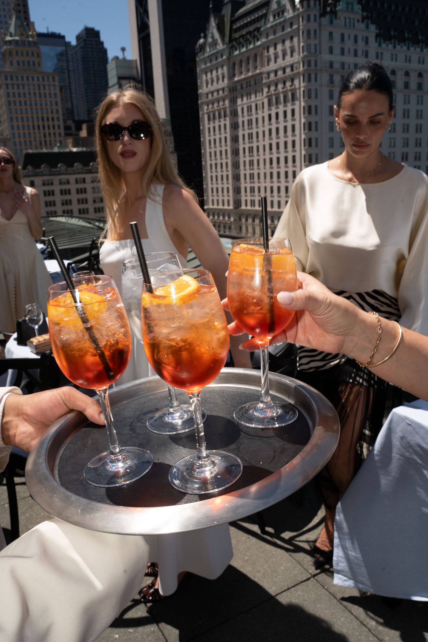 Server presenting tray of Aperol Spritz cocktails to women on a rooftop bar with NYC skyline views.
