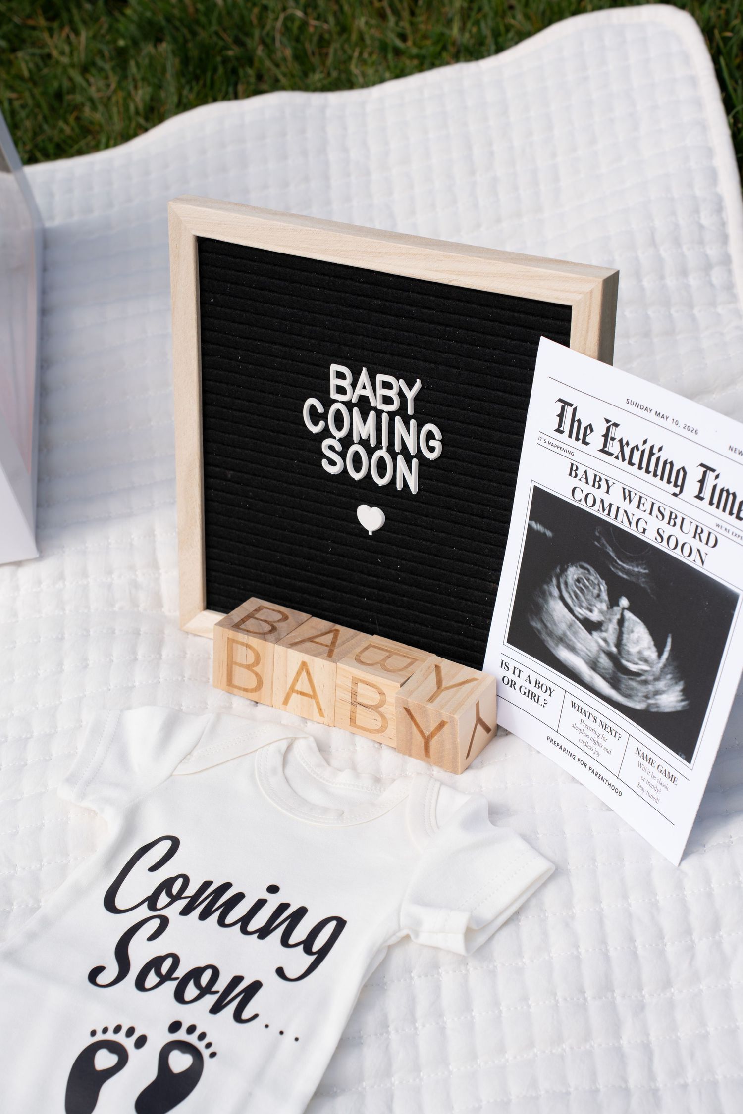 Baby announcement display with letter board, wooden blocks, ultrasound photo, newspaper, and onesie on white blanket.