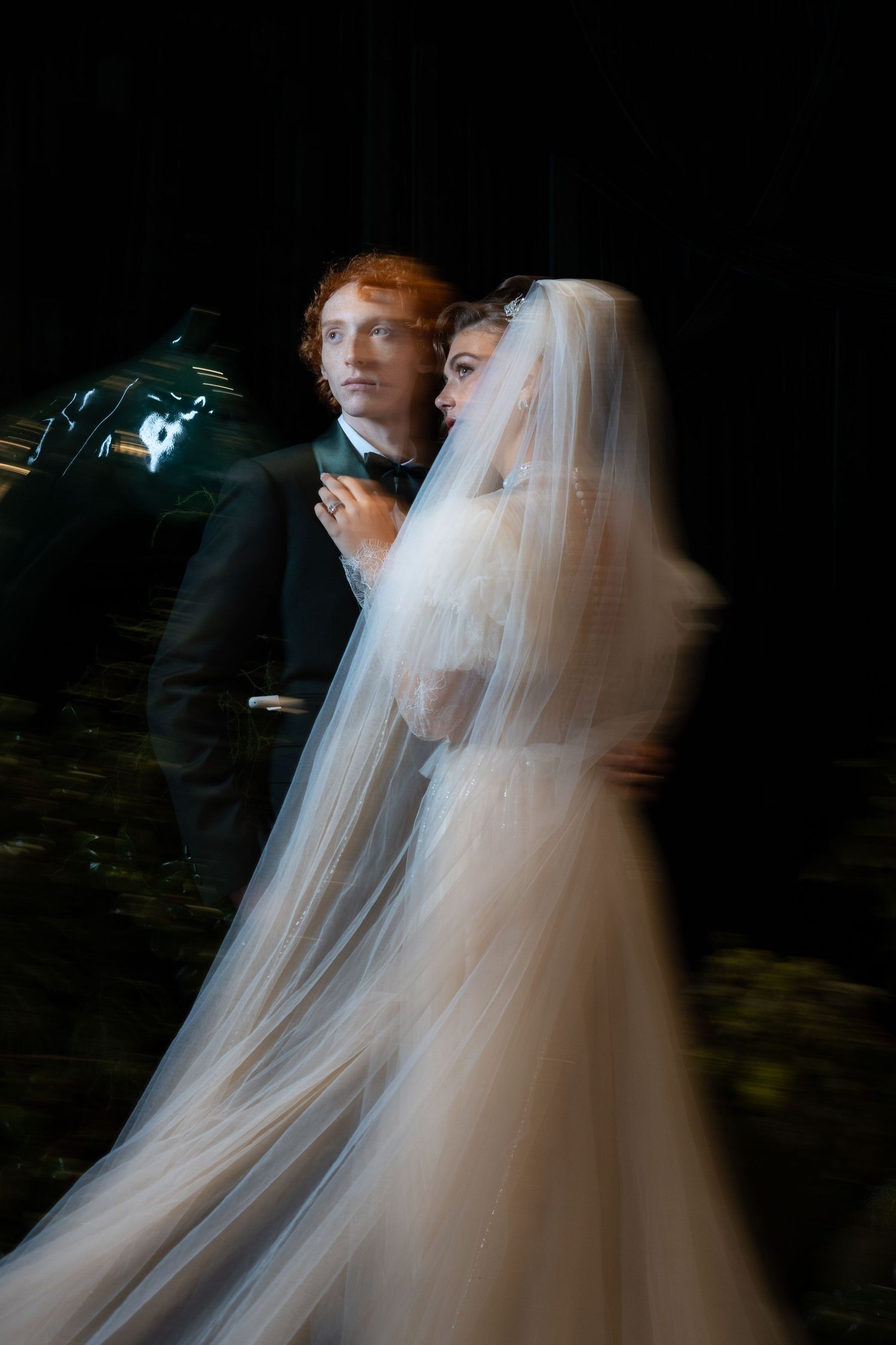 Motion-blur artistic wedding photo of bride in white tulle gown and red-haired groom embracing in dark setting.
