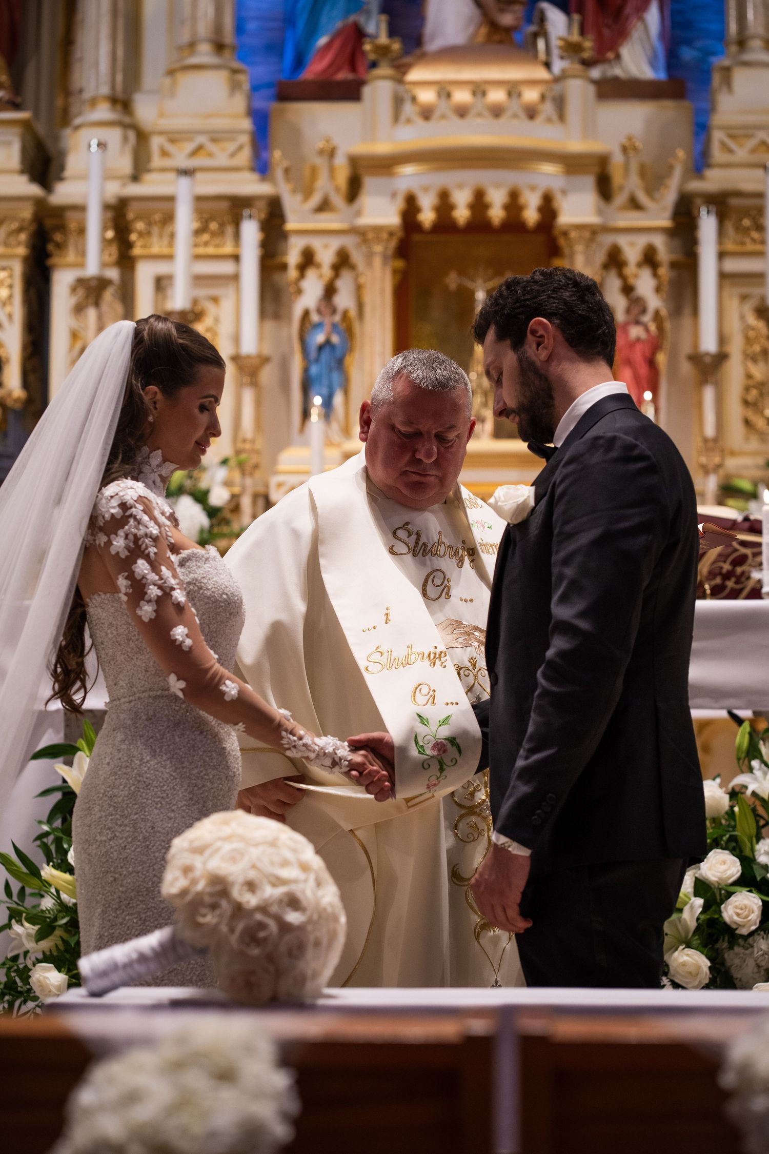 Catholic priest joins hands of bride and groom during solemn wedding blessing at ornate church altar.