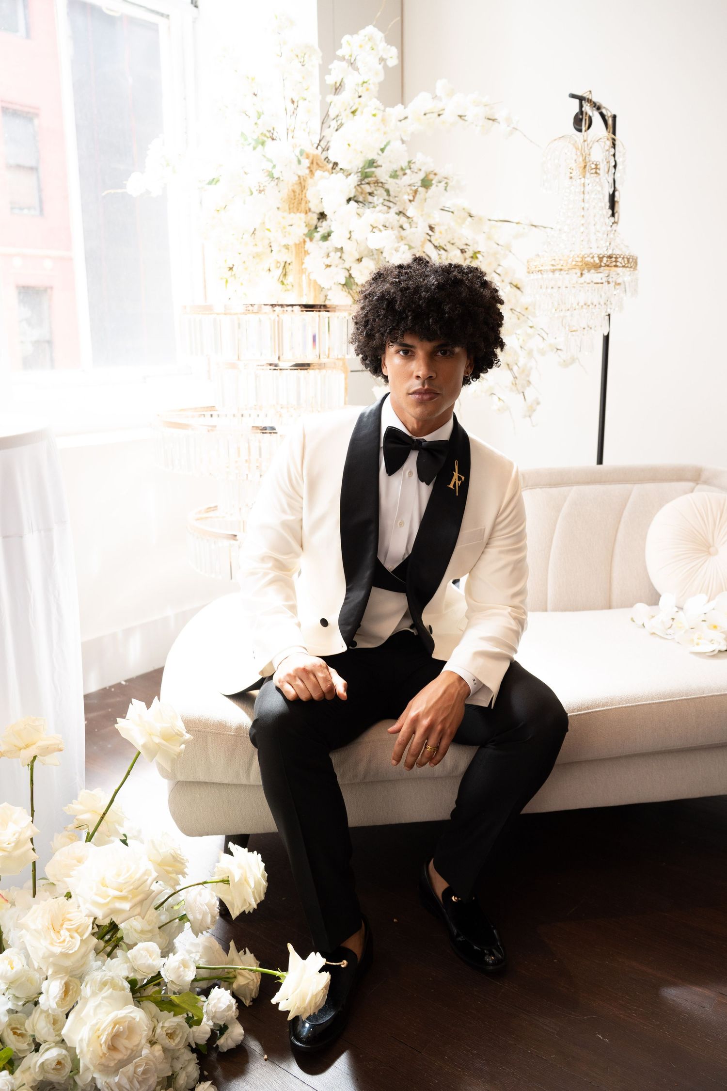 Dapper groom in white tuxedo sits on ivory sofa surrounded by white roses in bright wedding venue.