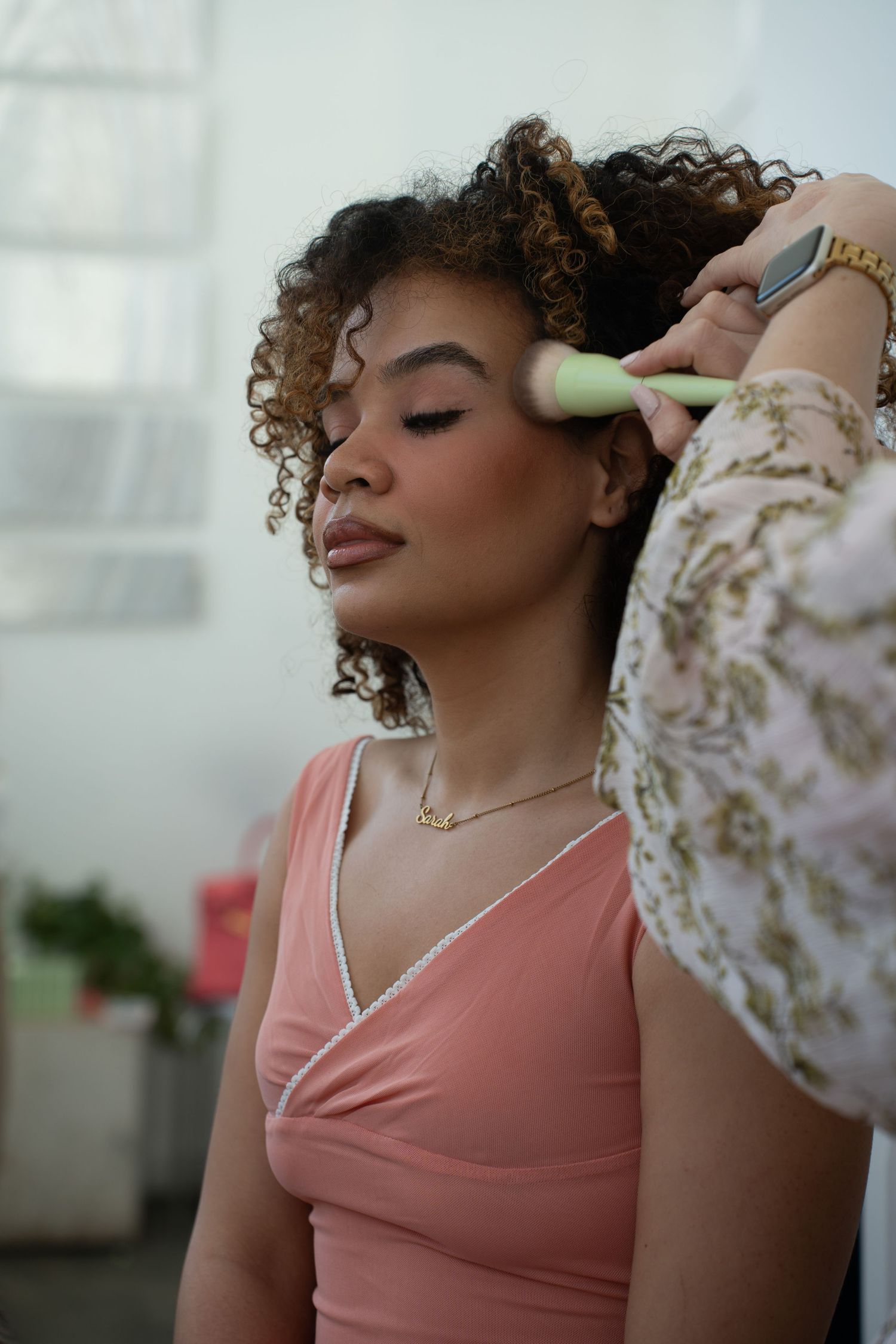 A makeup artist brushes powder on a woman with curly hair in a pink top, eyes closed.