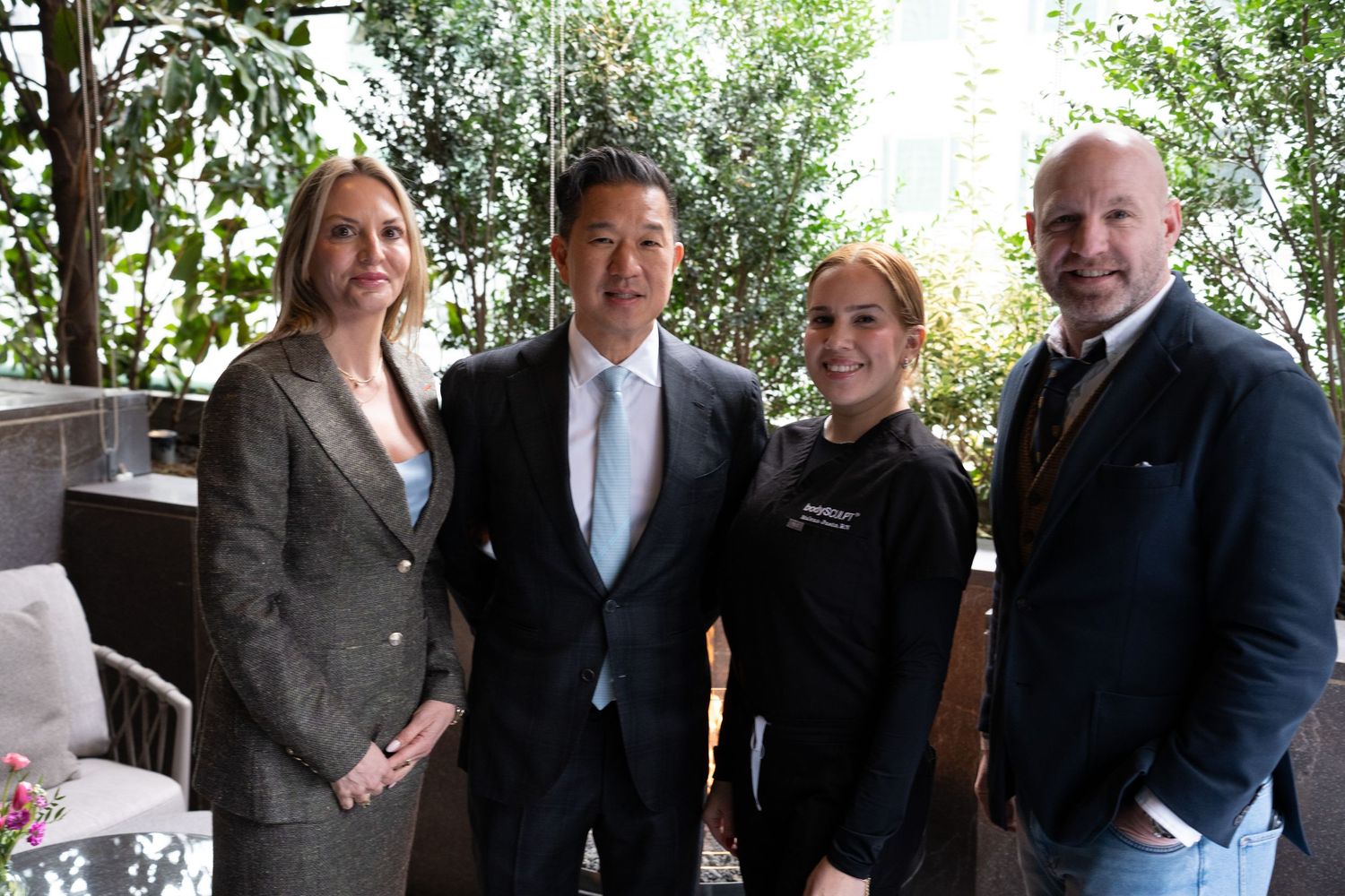 Four professionals posing outdoors at a medical aesthetics event with green foliage.