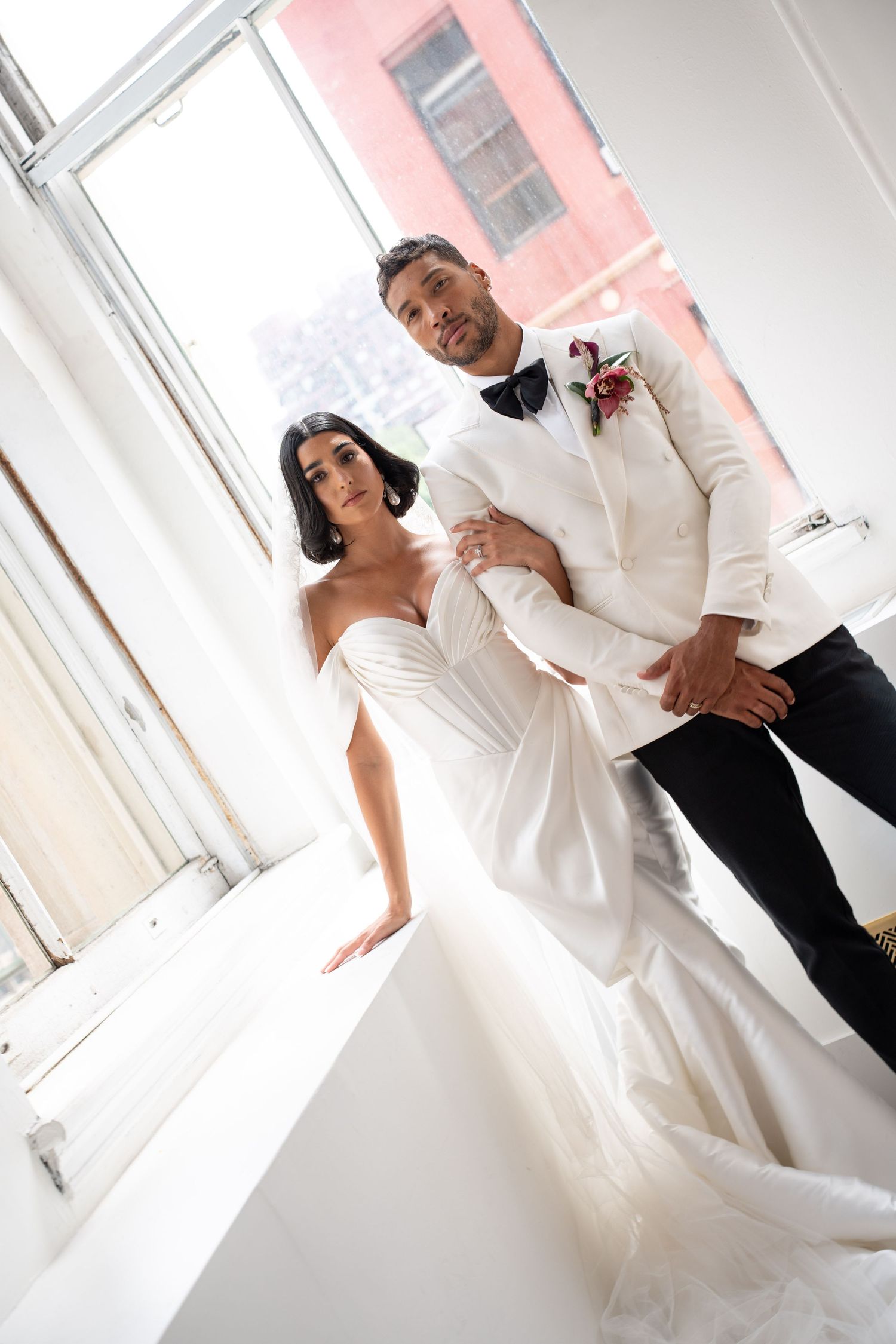 Bride and groom in white wedding attire standing together by bright window with city view.