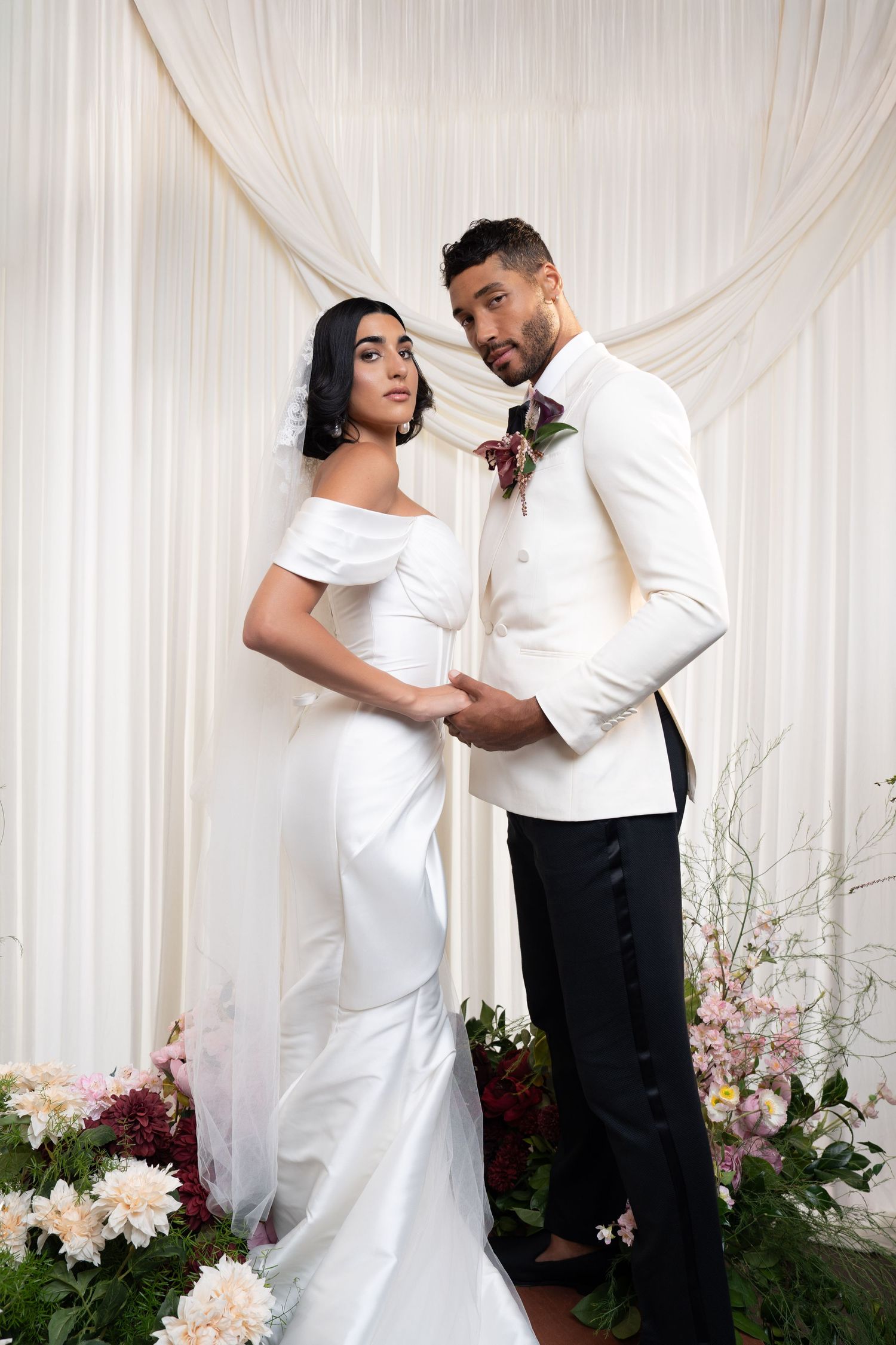 Bride and groom in white wedding attire hold hands surrounded by lush floral arrangements indoors.