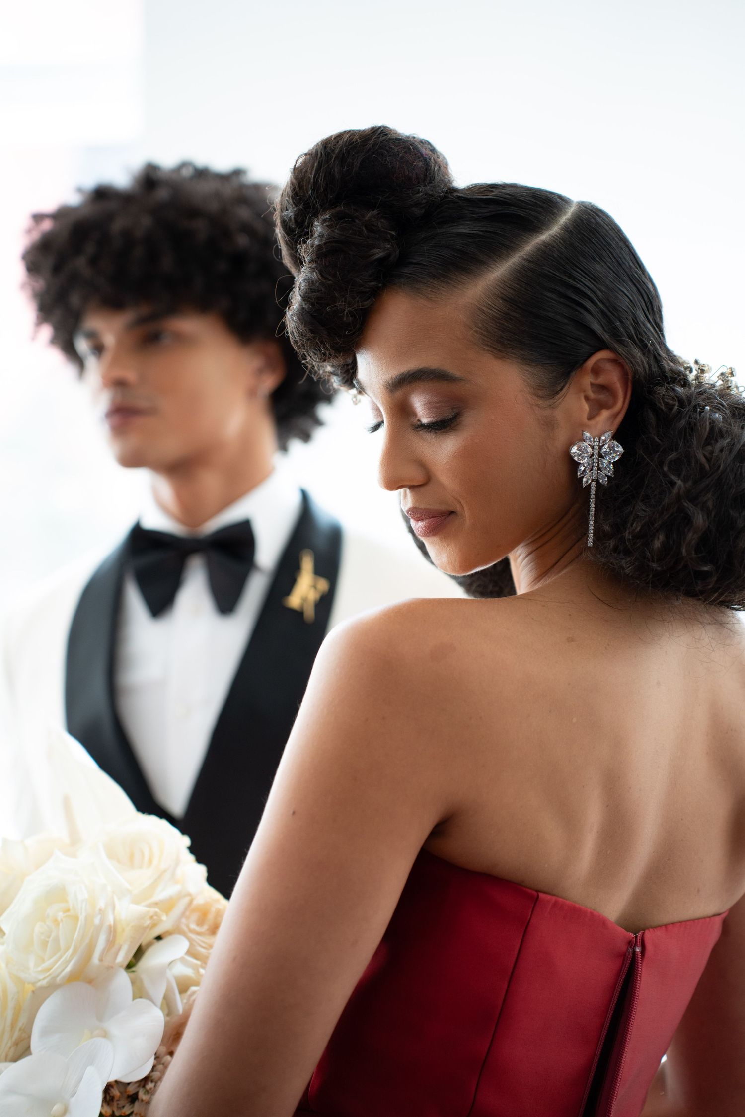 Woman in red strapless gown with ornate updo and drop earrings looks down while man in tuxedo stands behind her.