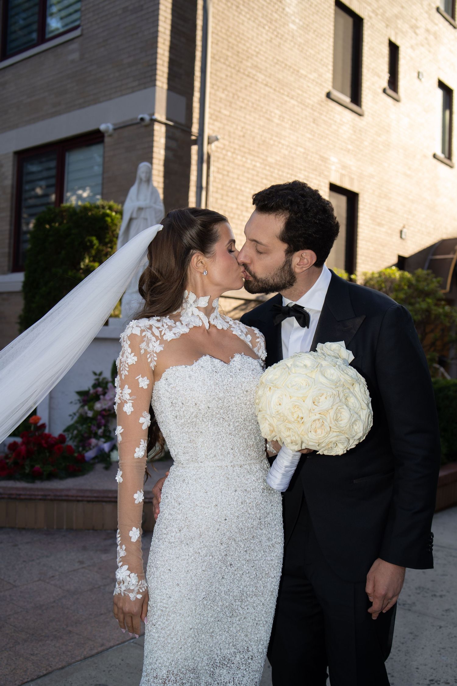 Newlyweds share a kiss outdoors, bride in beaded lace gown holding white rose bouquet.